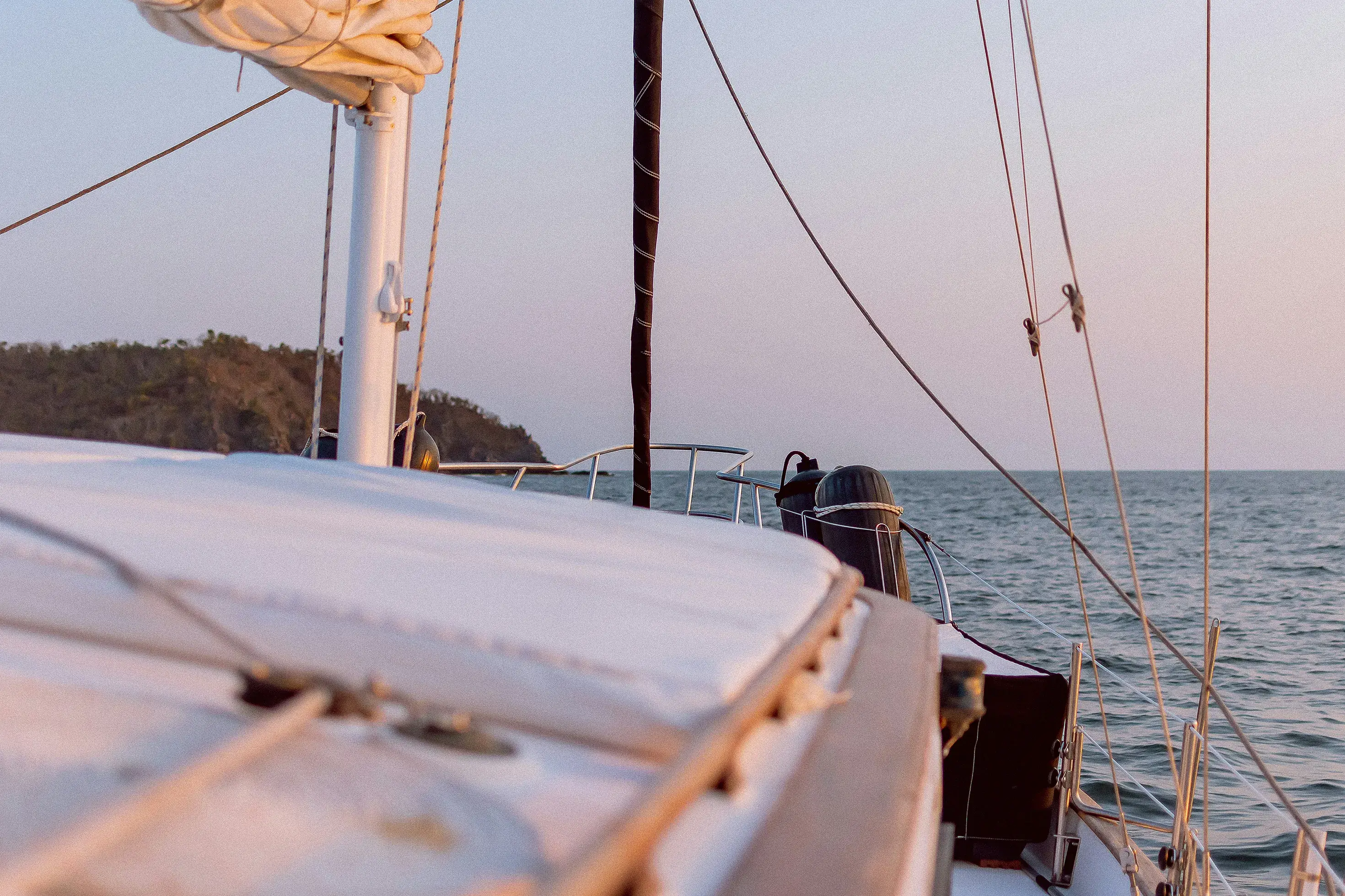 Peaceful view from a sailboat deck sailing on the ocean at sunset, with mast, ropes, and a distant coastline under soft golden light.