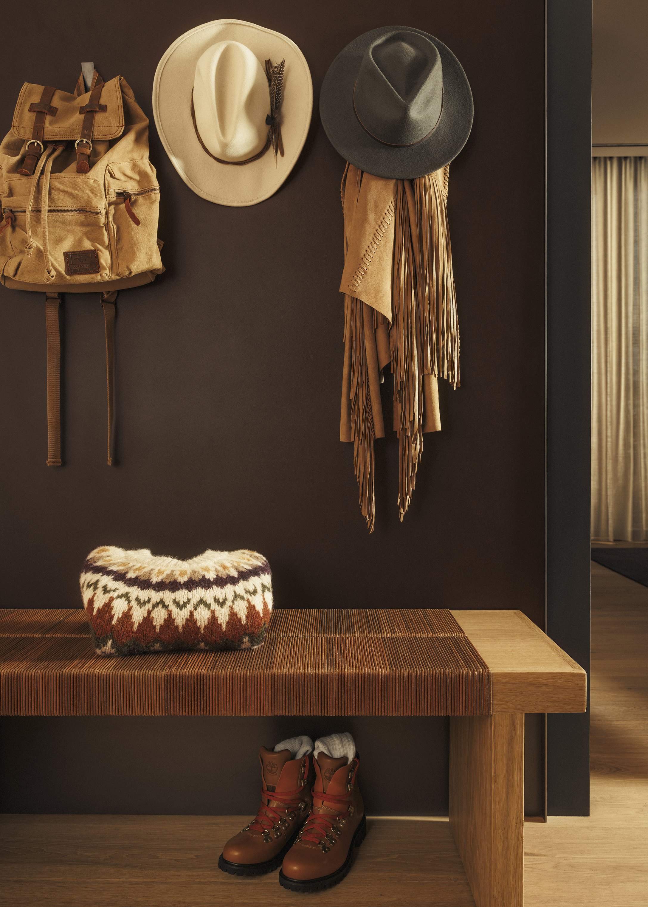 A wall featuring two hats, a scarf and a rucksack hanging above a bench with a folded jumper on top and boots below