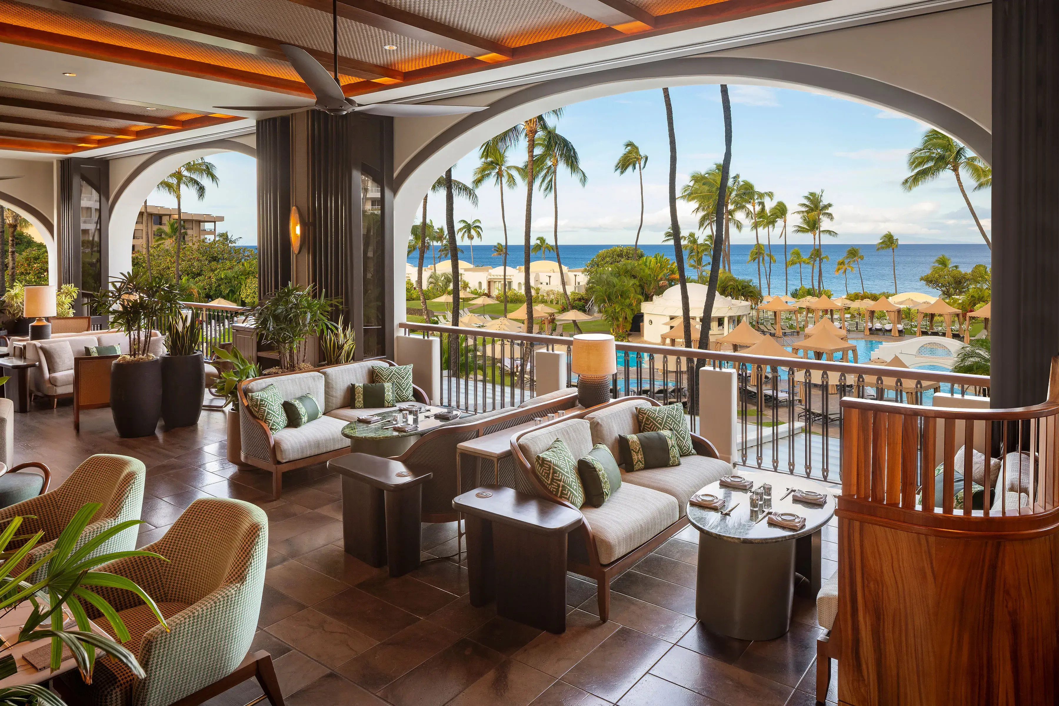 The comfy terrace area of PILINA Restaurant at Fairmont Kea Lani featuring wooden furniture with green decor beside large archways overlooking the resort and ocean