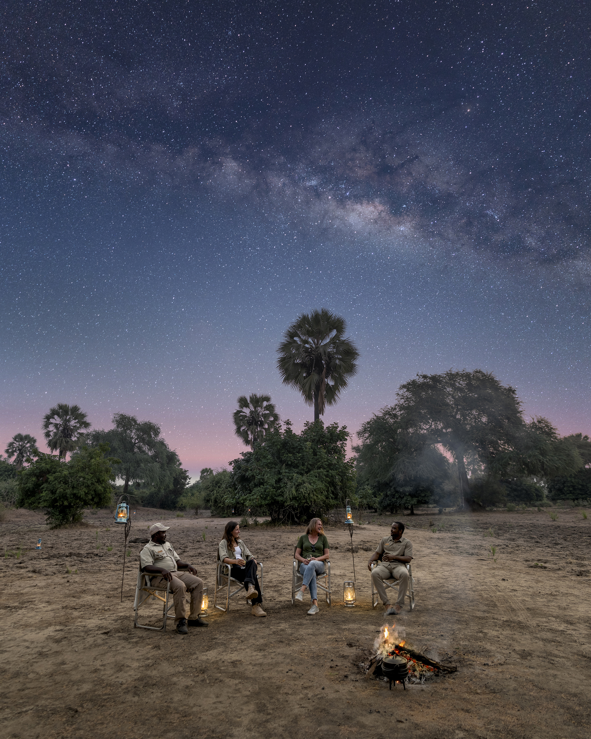 Four people sitting around a campfire under a starry night sky with the Milky Way visible, surrounded by trees in a natural setting.
