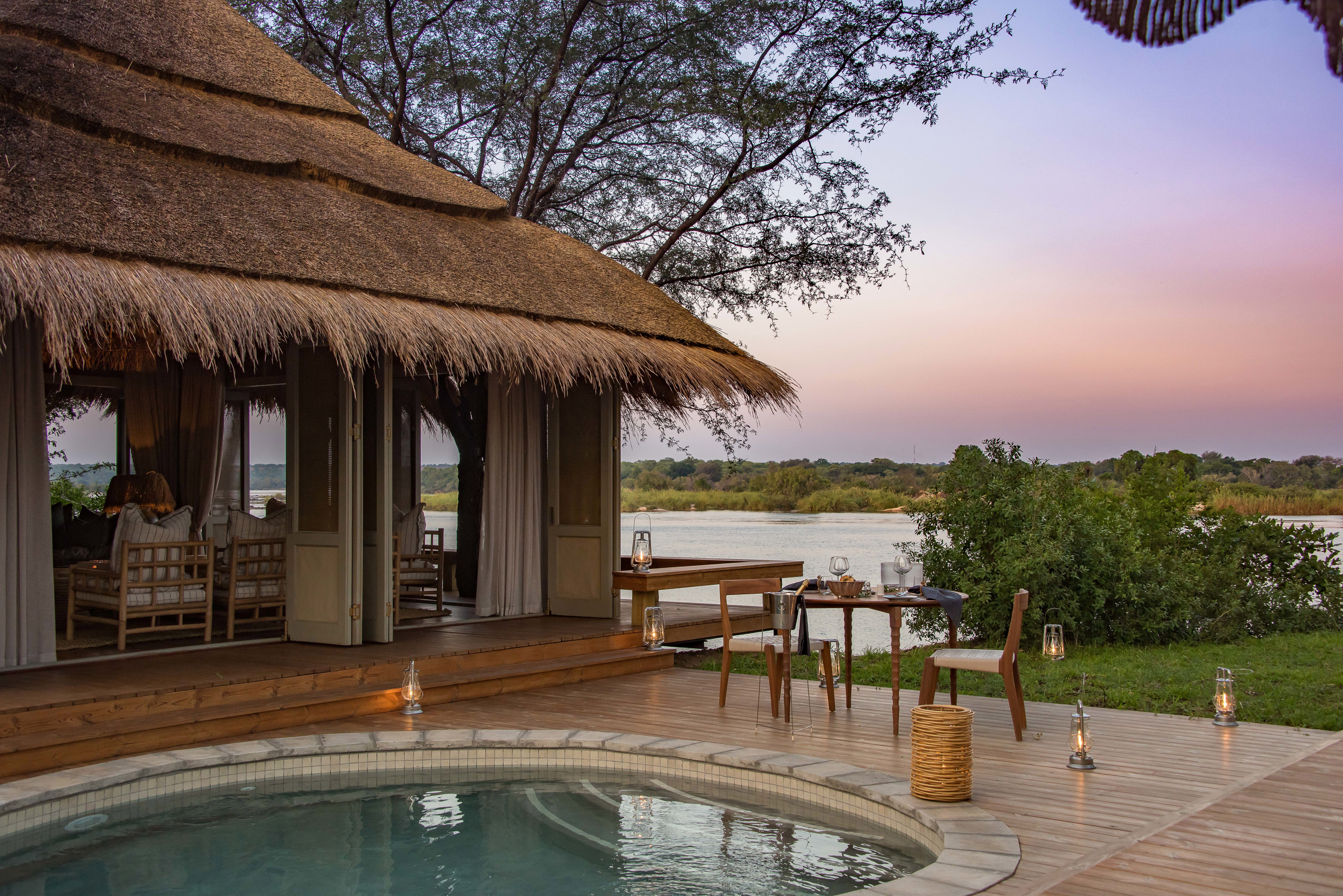 An private dinner table set beside an outdoor pool next to a thatched building opposite the Zambezi river