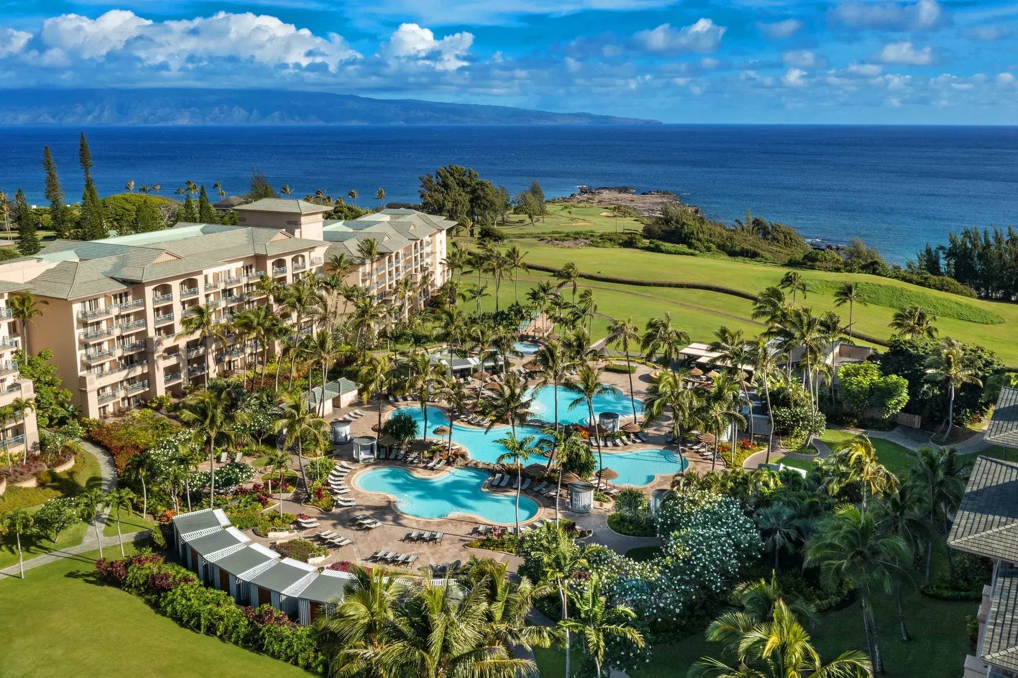 An aerial view of The Ritz-Carlton Maui, Kapalua resort featuring three swimming pools, palms trees and the hotel exterior nestled among a well kepy golf course with the ocean behind