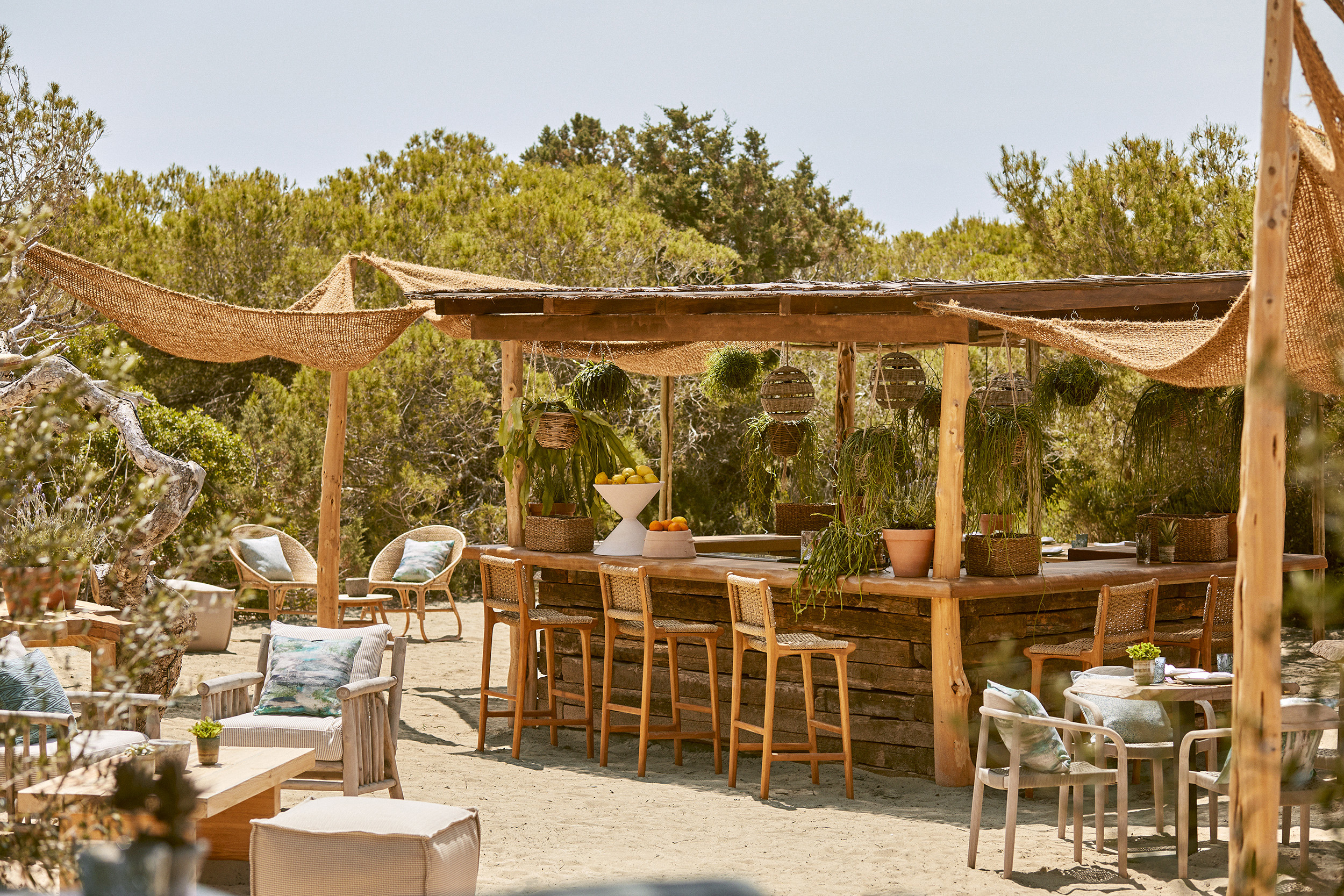A relaxed boho garden kitchen area with wooden furniture decorated in natural soft furnishings surrounded by plants beneath a clear sky