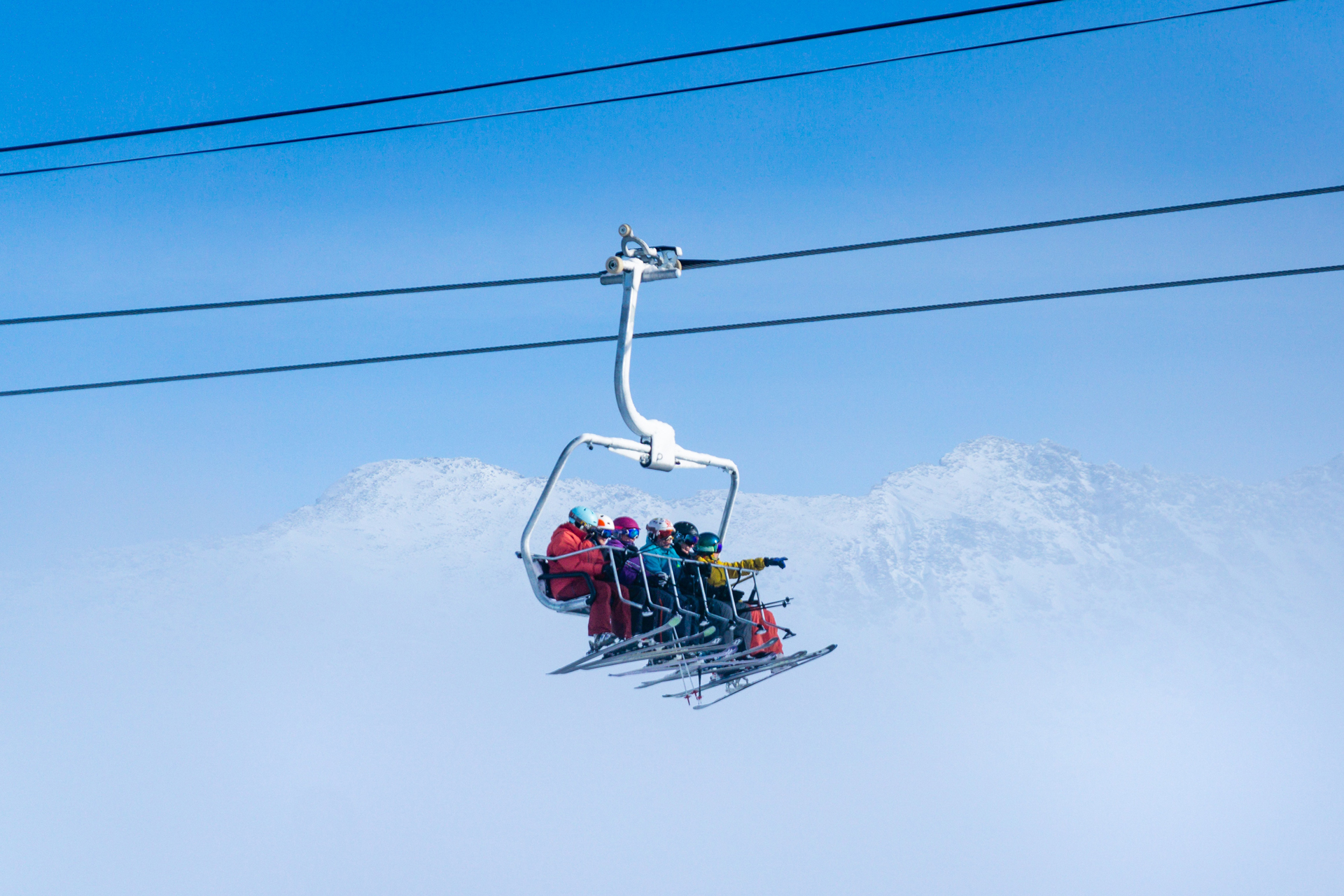 A group of skiers on a ski lift with a partially visible mountain range behind under a blue sky