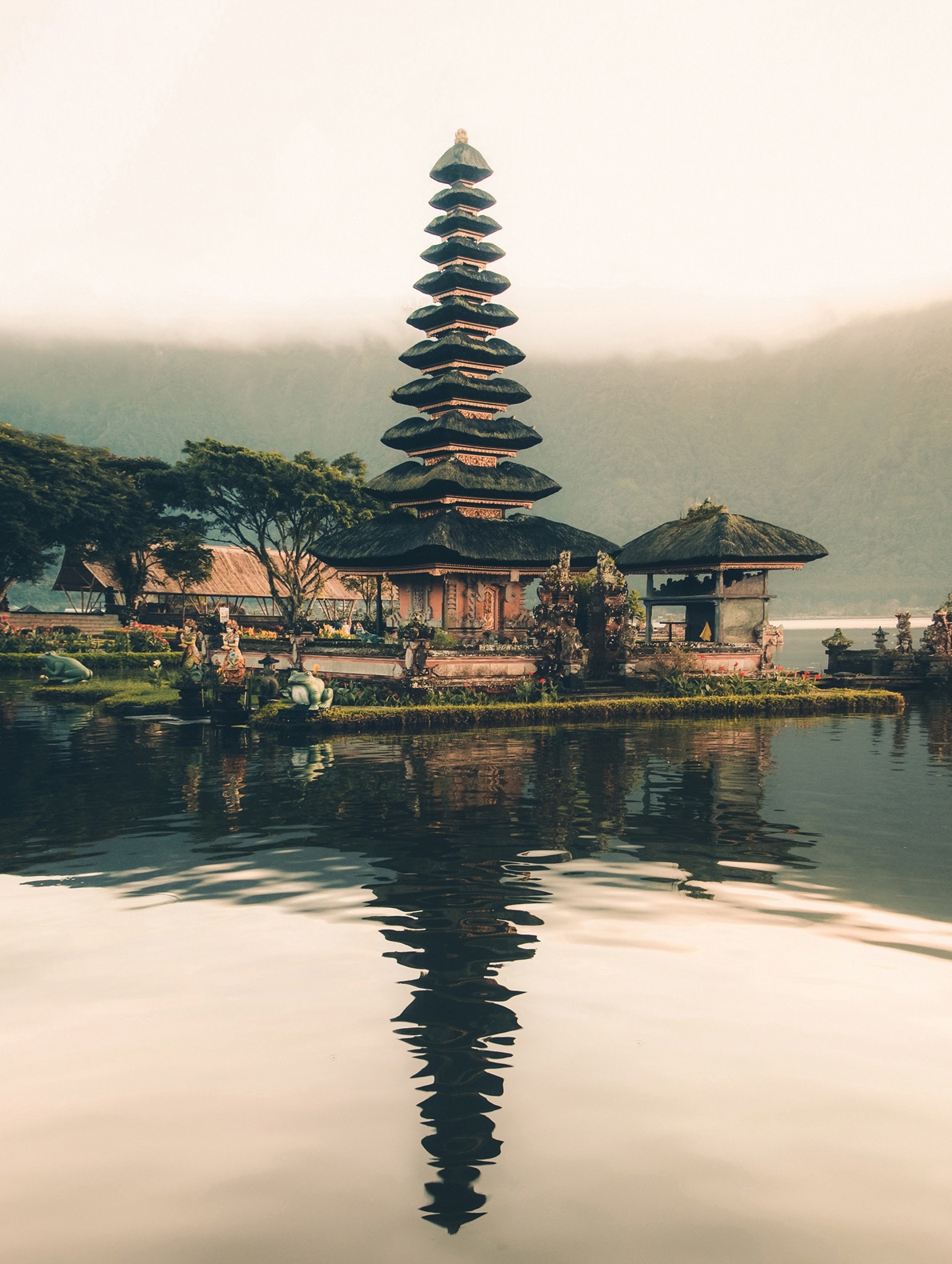 A multi-tiered pagoda-like temple is reflected in calm water, surrounded by greenery and misty mountains.
