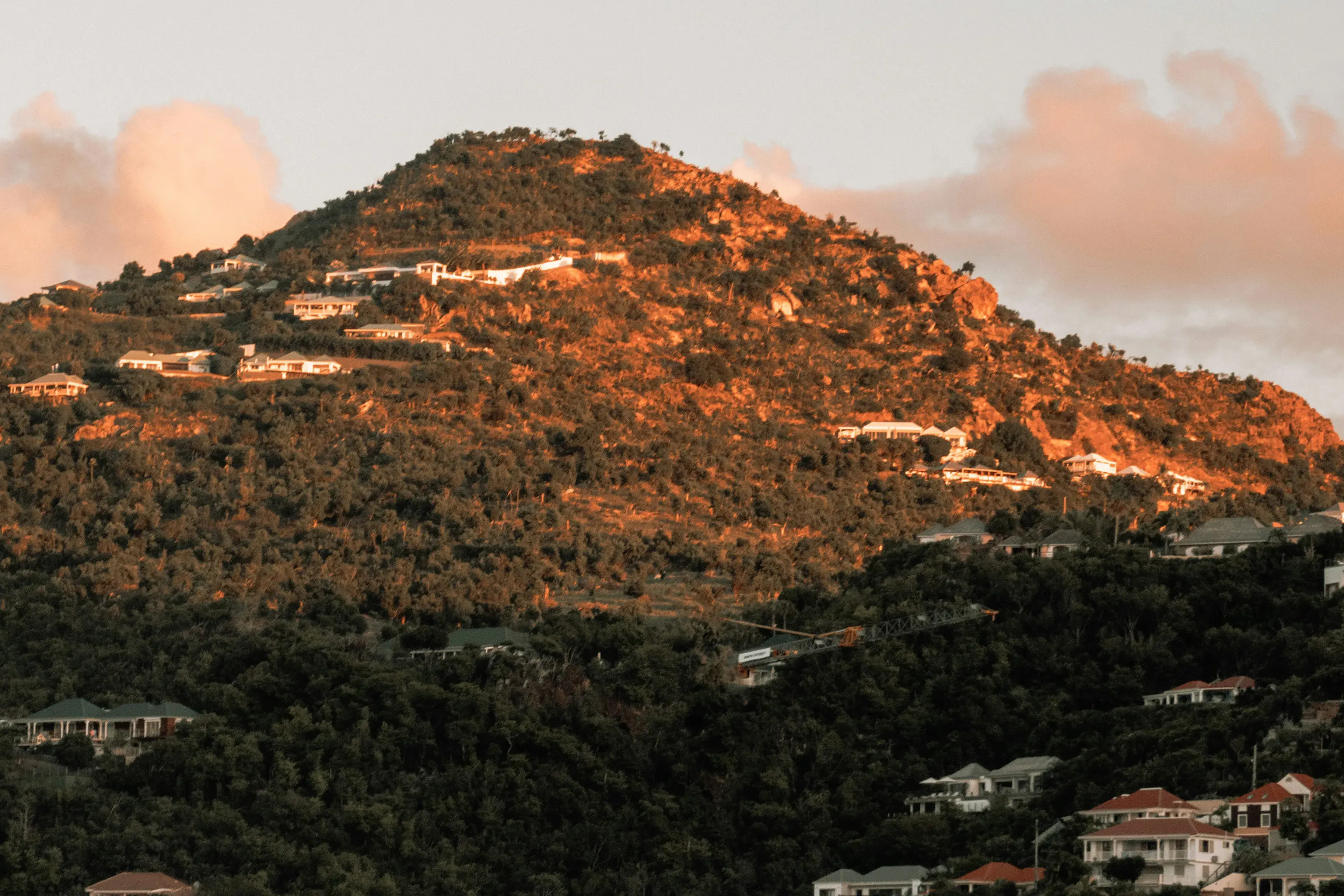 Sunlit hillside on St Barths dotted with scattered homes and lush greenery under a soft, pastel sky.
