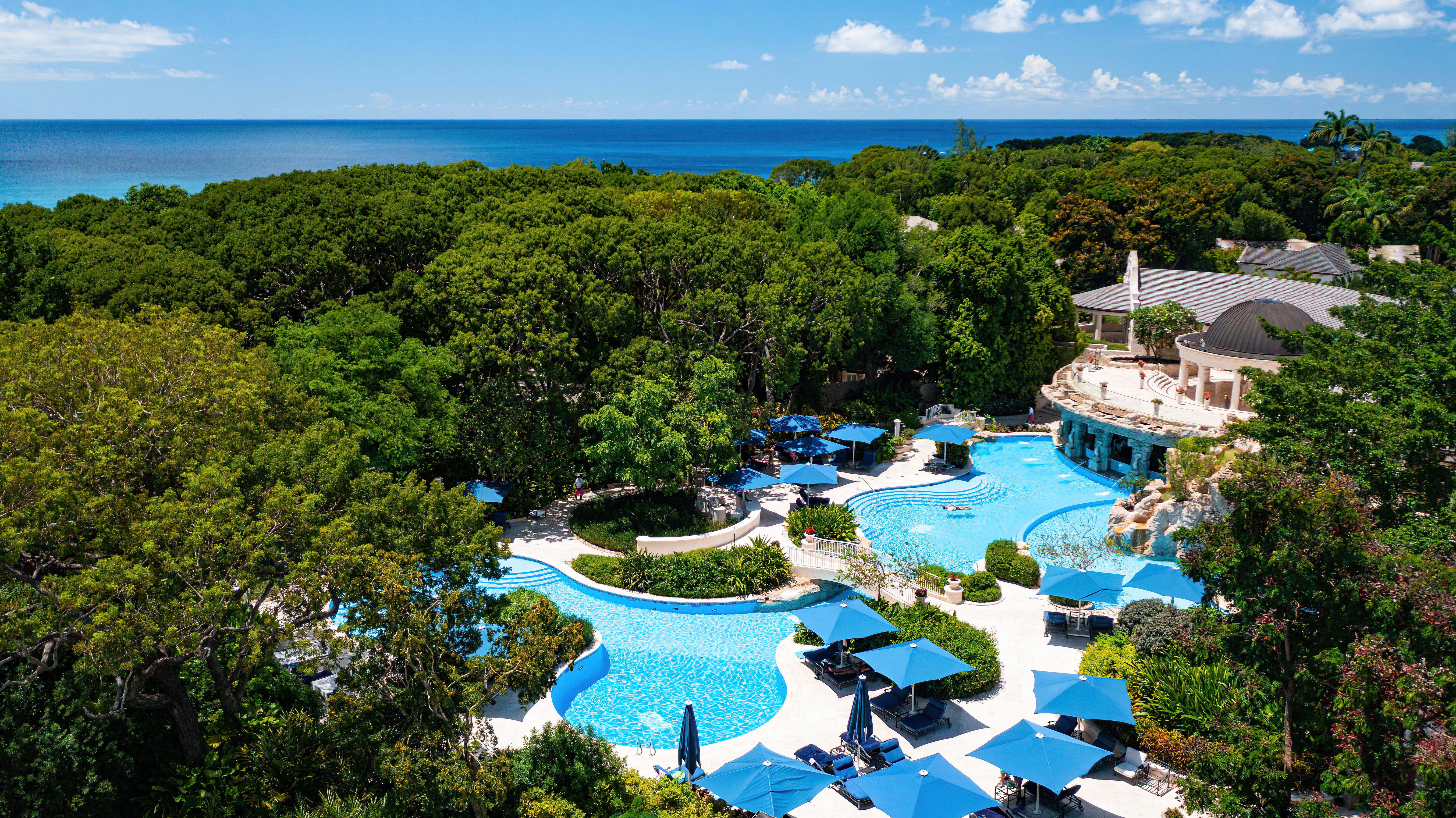 The opulent spa building at Sandy Lane with a large blue swimming pool surrounded by blue sun umbrellas within greenery