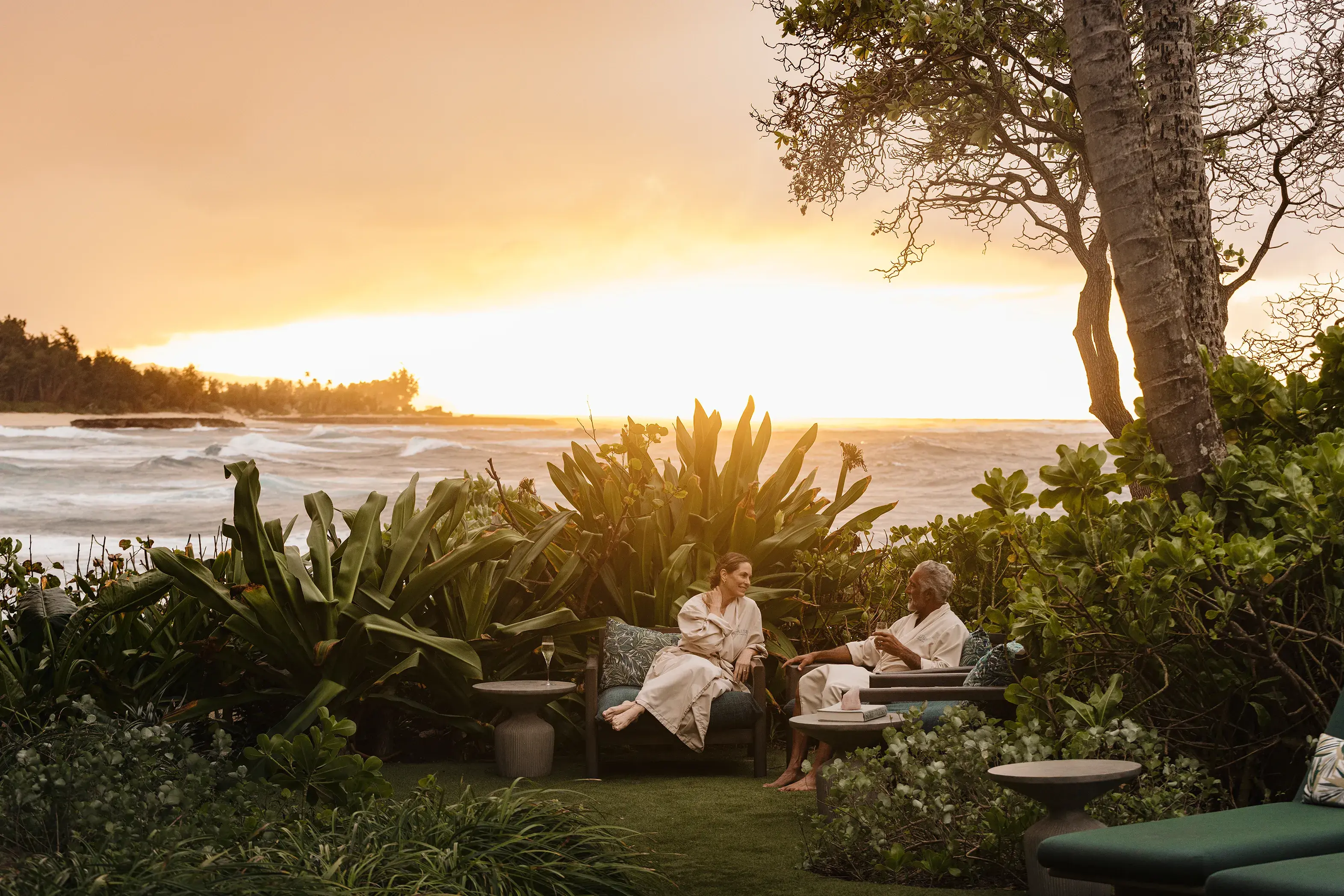 Two guests relaxing in the spa garden wearing fluffy white robes with the sun setting behind