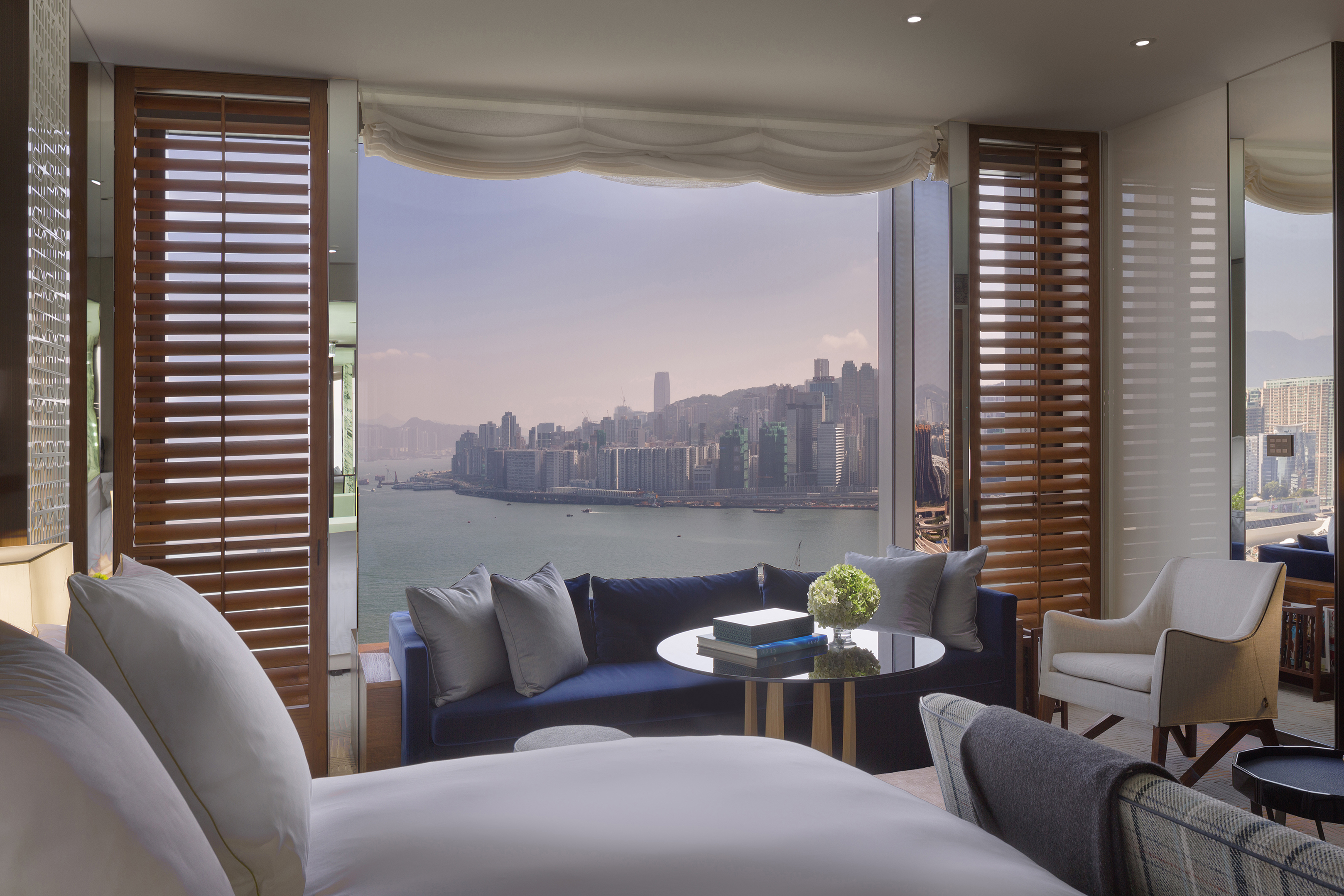 The bedroom of a Harbour View Room showing comfy furniture in front of wooden shutters framing a large window with city views