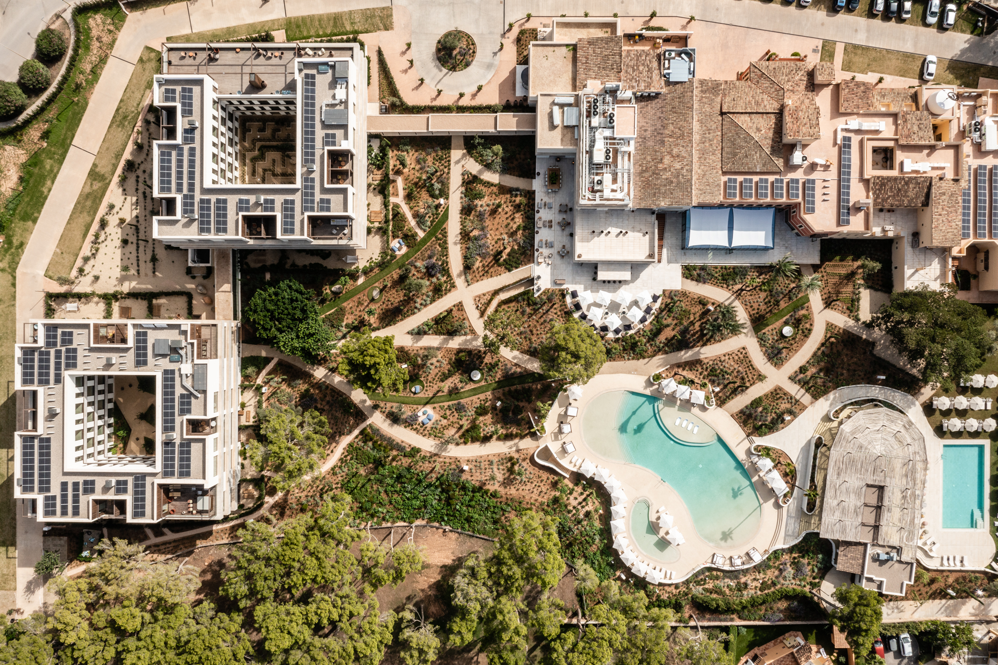 Aerial view of a landscaped property with geometric gardens, two swimming pools, and adjacent buildings.