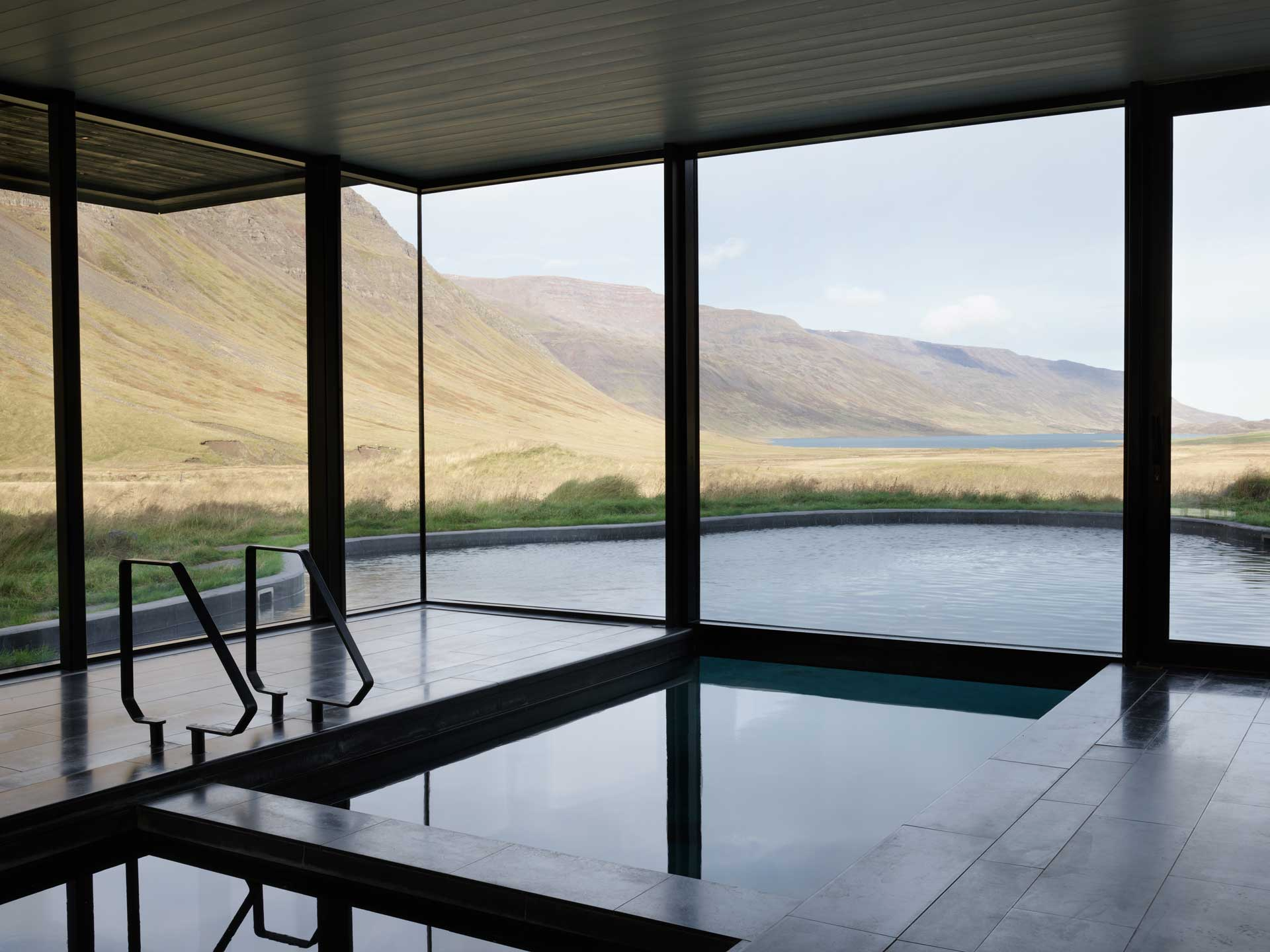 Minimalist indoor pool with floor‑to‑ceiling windows overlooking an expansive valley and distant water.