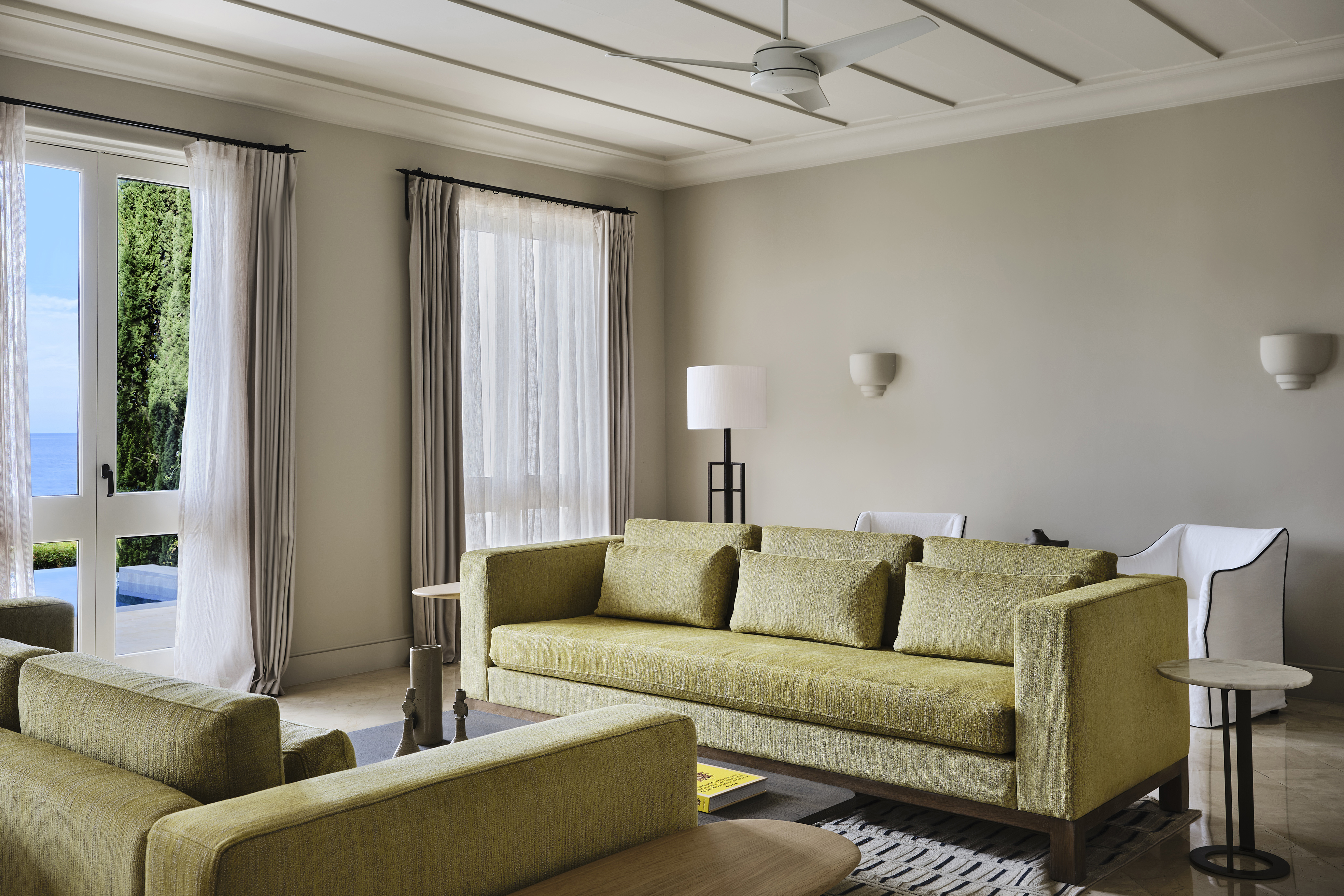 The living room of the Alcyone Residence featuring soft green sofas in a pale room surrounded by sleek furniture with french doors leading to a balcony