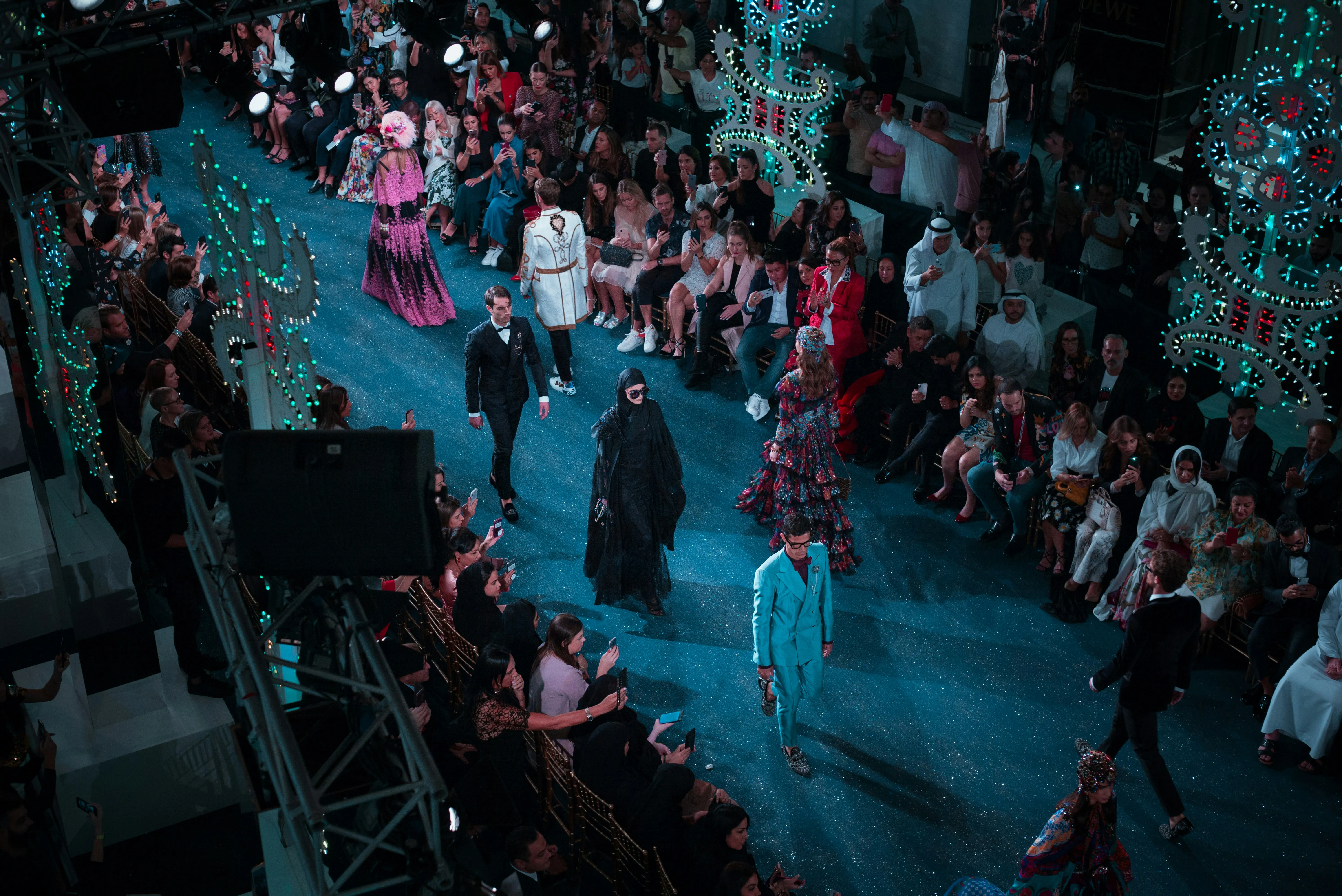 Fashion runway show with models wearing vibrant designer outfits walking on a glittering blue catwalk surrounded by seated audience and ornate decorative lights.