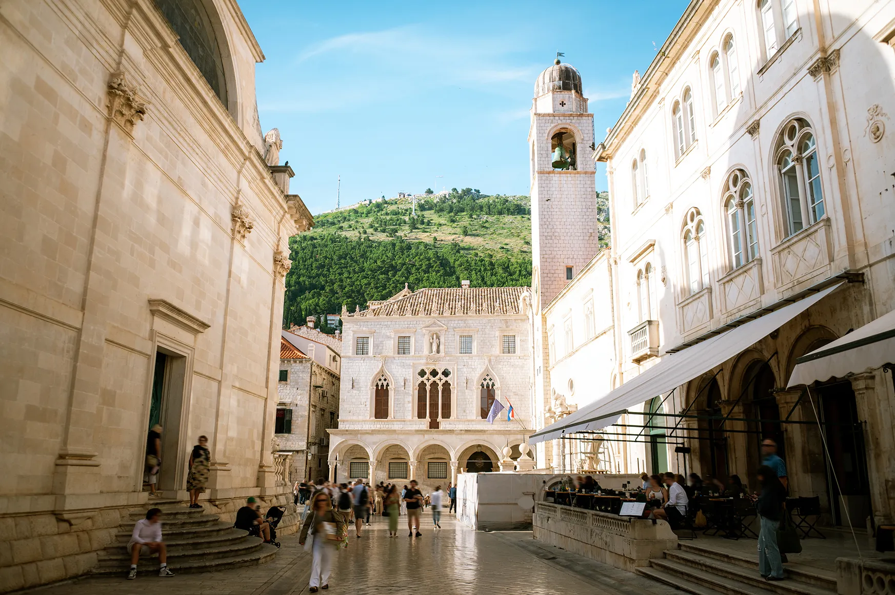 Historic stone square with bell tower and pedestrians, framed by ornate buildings and a hillside backdrop.