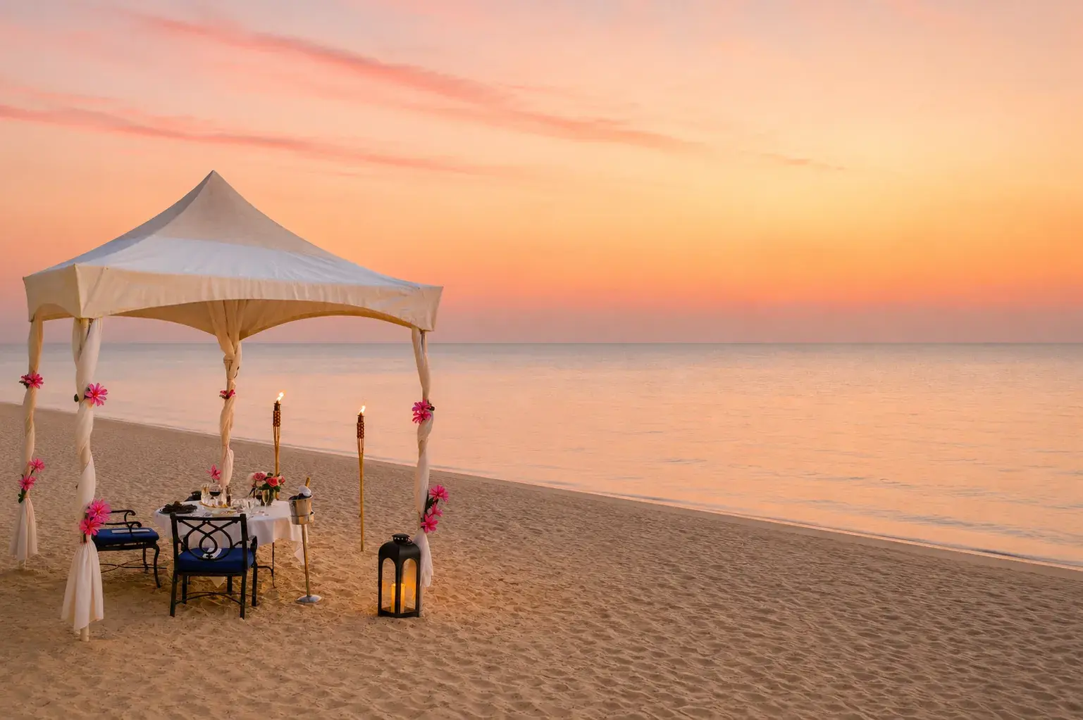 A romantic private dining set up on the beach at sunset featuring an elegant table and torches set underneath a white tent with pink flowers