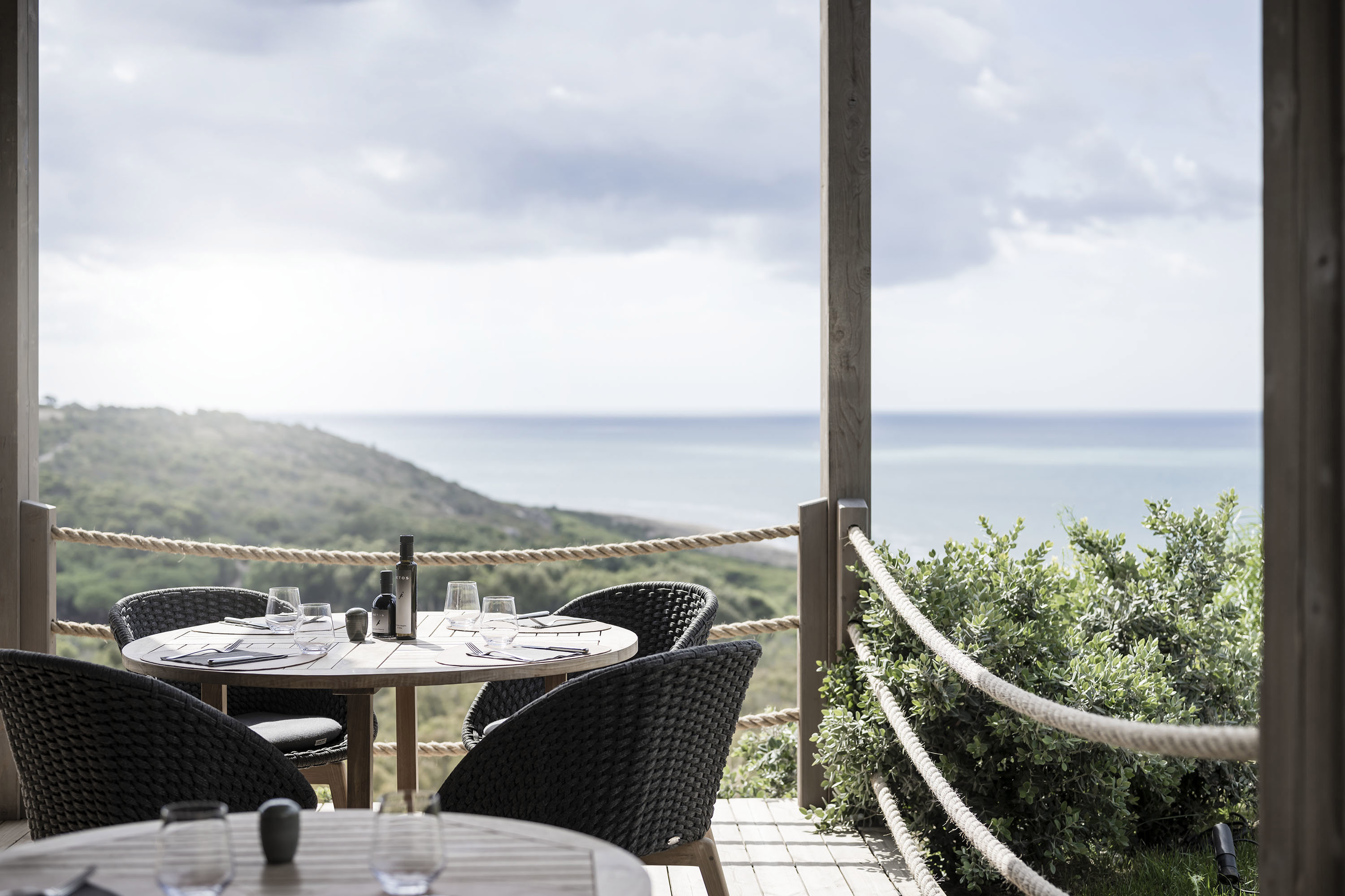 A covered terrace on a green hillside beside the sea with circular tables and dark rattan chairs set for lunch