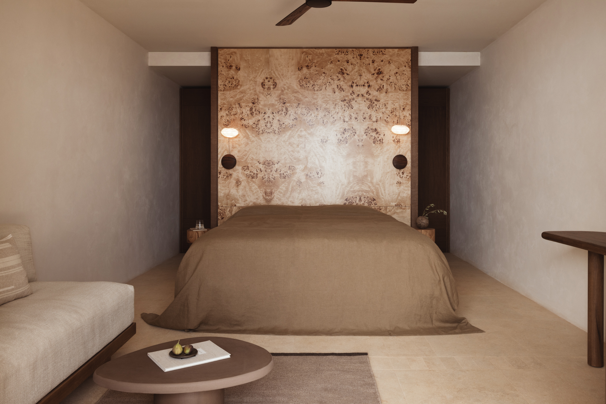 A bed with brown linens set against a patterned partition wall with a glimpse of a coffee table and sofa in front