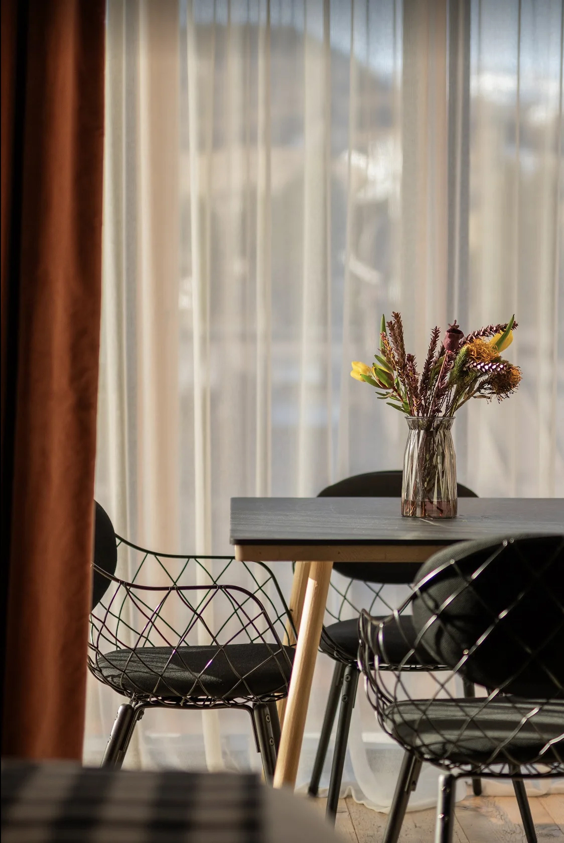A vase of dried flowers atop a dining table with two metal chair and sheer curtains