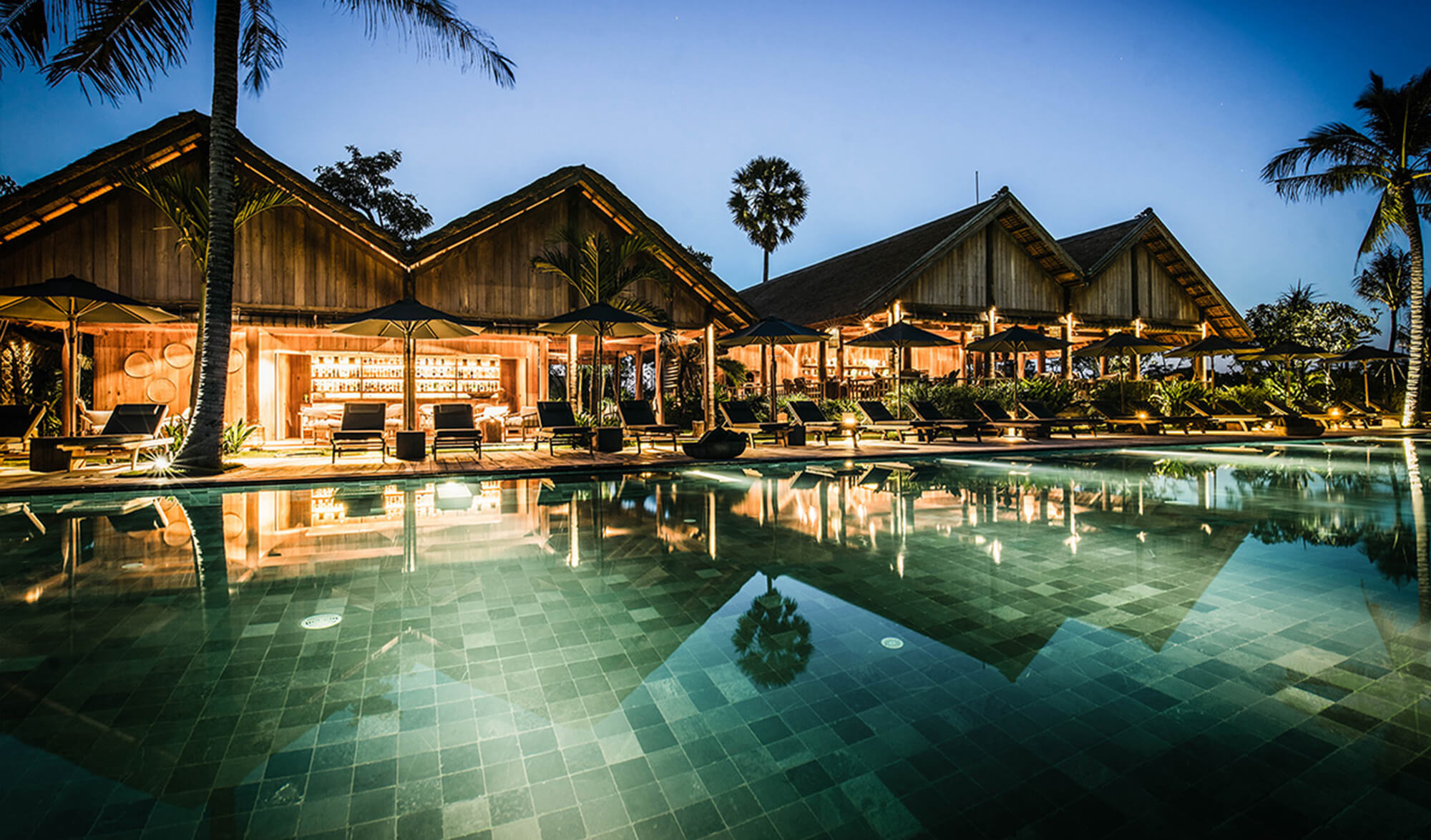 Luxurious resort with thatched-roof buildings around a large illuminated pool at twilight