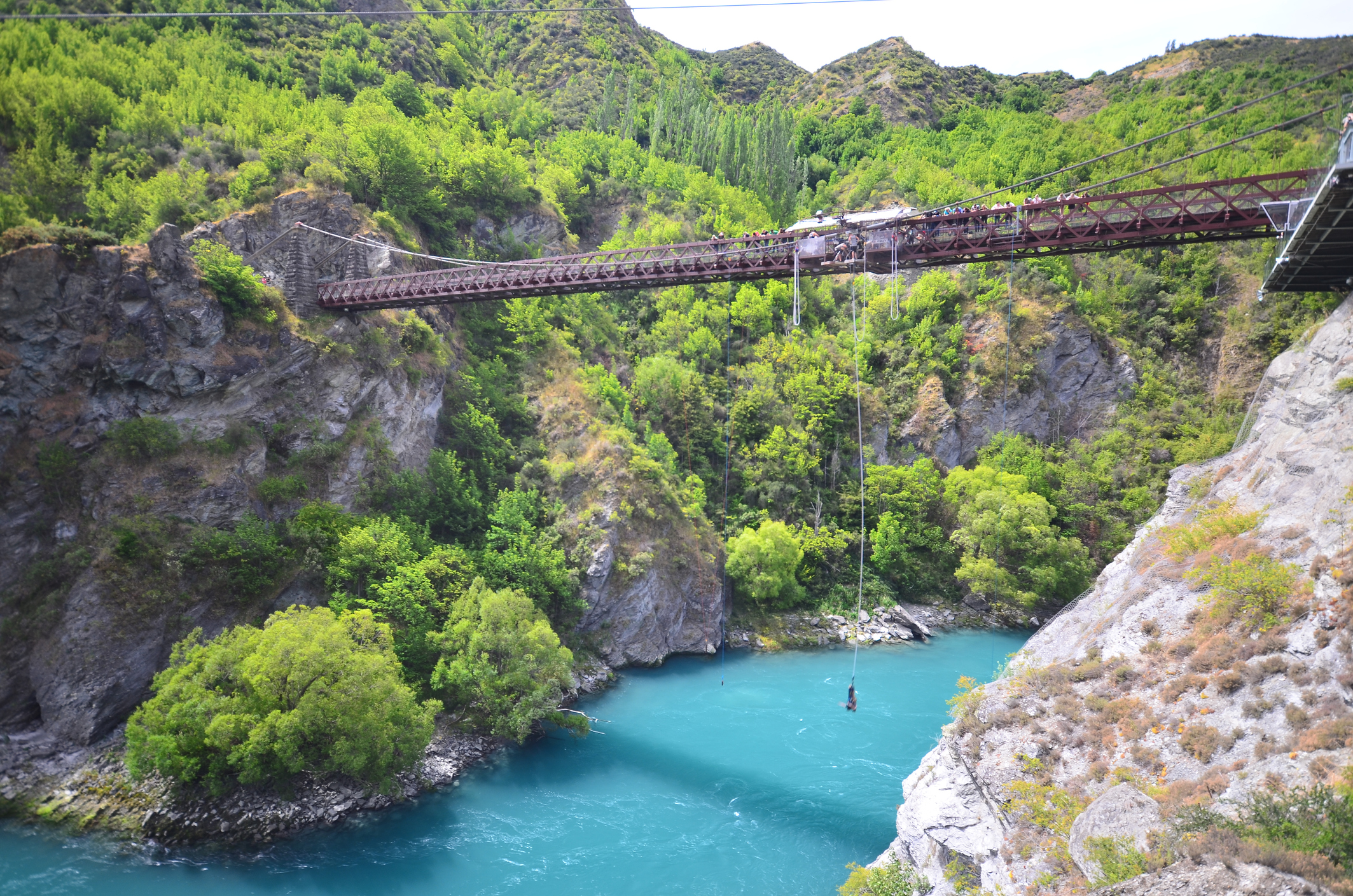 A person is bungee jumping off a bridge over a turquoise river surrounded by lush green trees and rocky cliffs