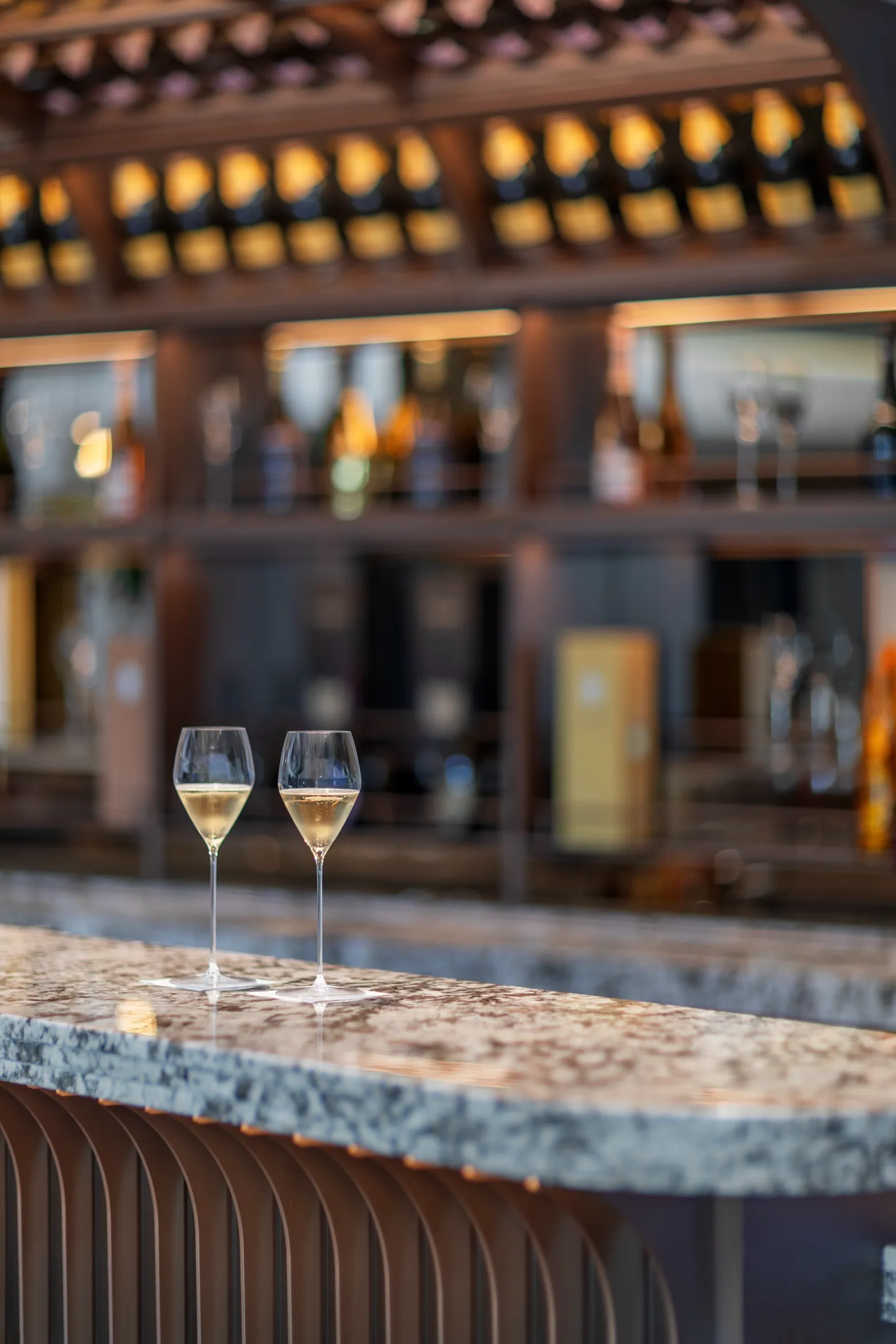 Two champagne glasses on a marble bar counter with shelves of bottles in the background.