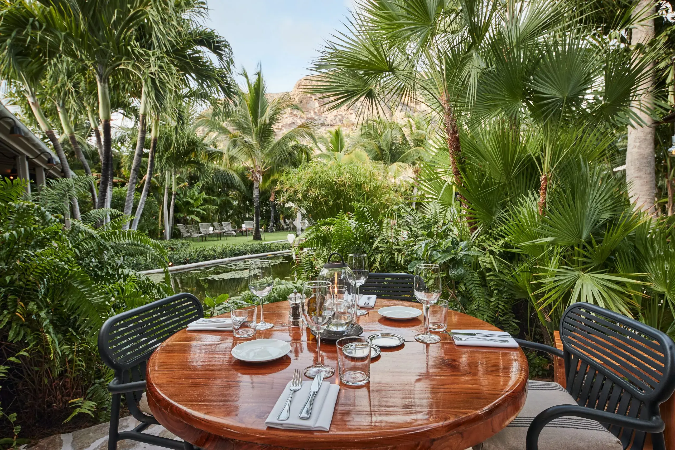 Outdoor dining table set for a meal surrounded by lush tropical gardens and palm trees beside a tranquil pond.