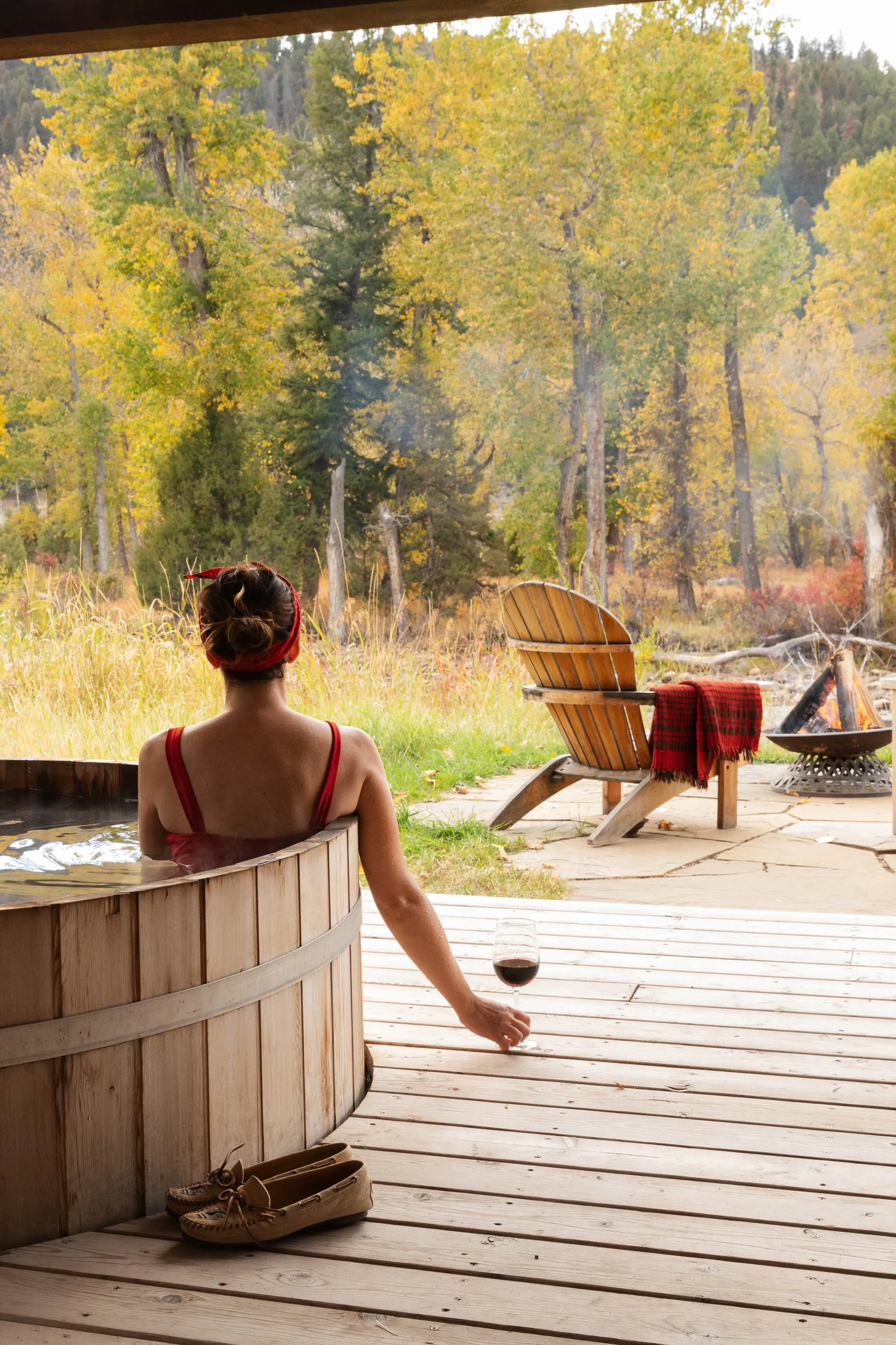 Deck of Sweet Grass canvas cabin at The Ranch at Rock Creek in Montana featuring a woman in a wooden hot tub holding a glass of wine, with a wooden chair and small fire set in front of a copse of trees
