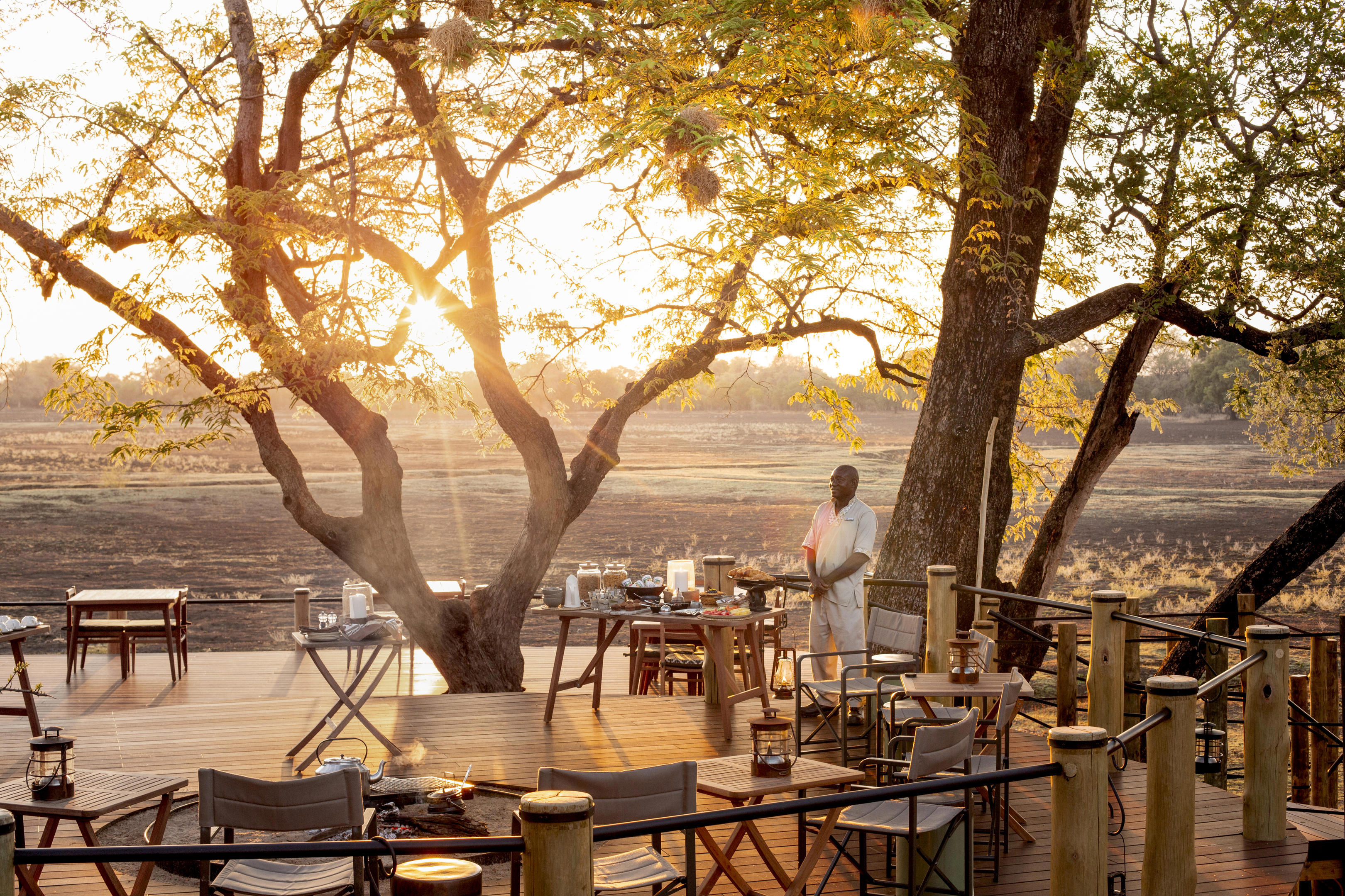 Outdoor dining area at Puku Ridge Camp with a wooden deck, tables, and chairs set beneath large trees during sunset