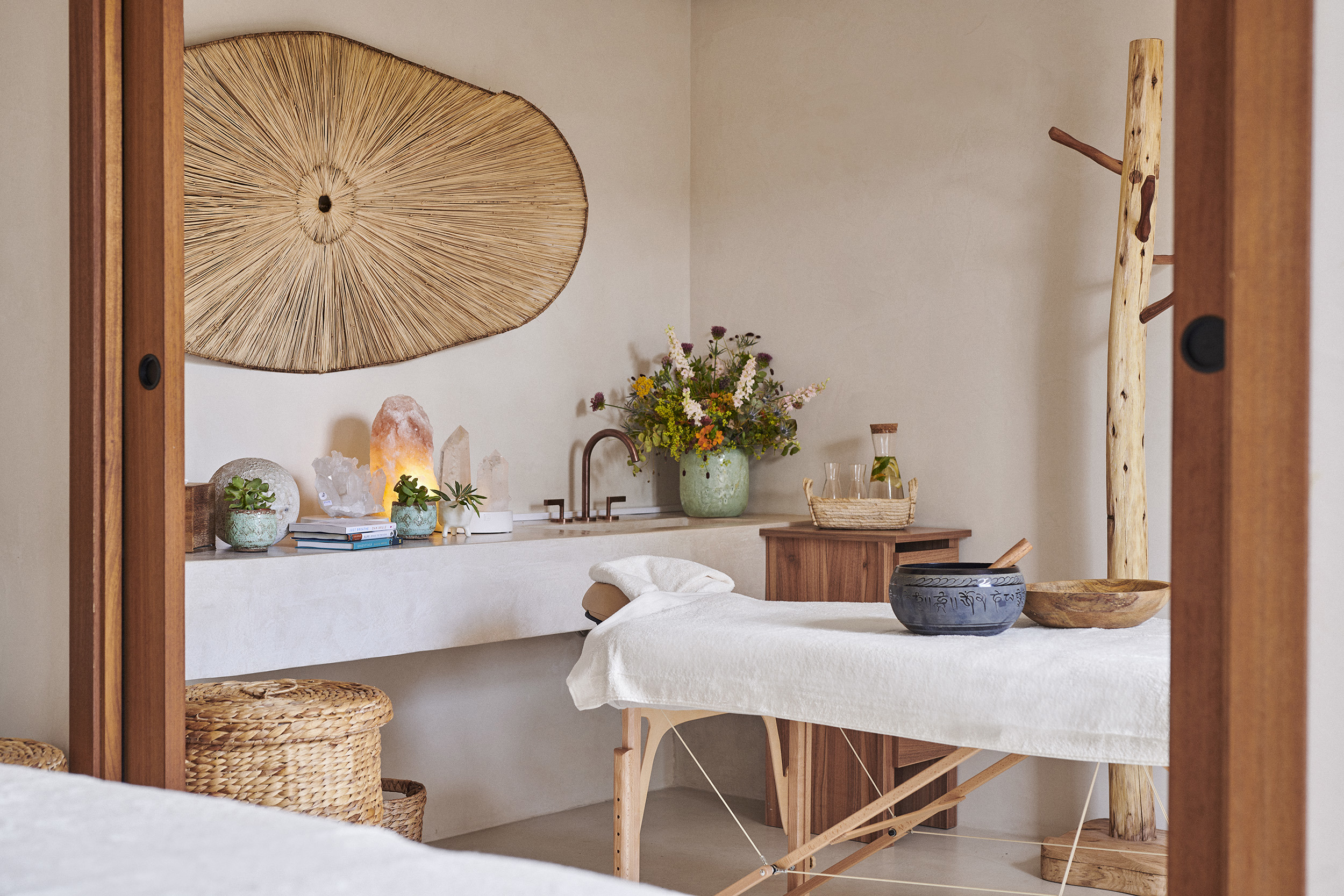 A spa treatment room at Teranka featuring a massage bed set with bowls in front of a sink area lined with books, crystals and a vase with flowers beneath straw artwork