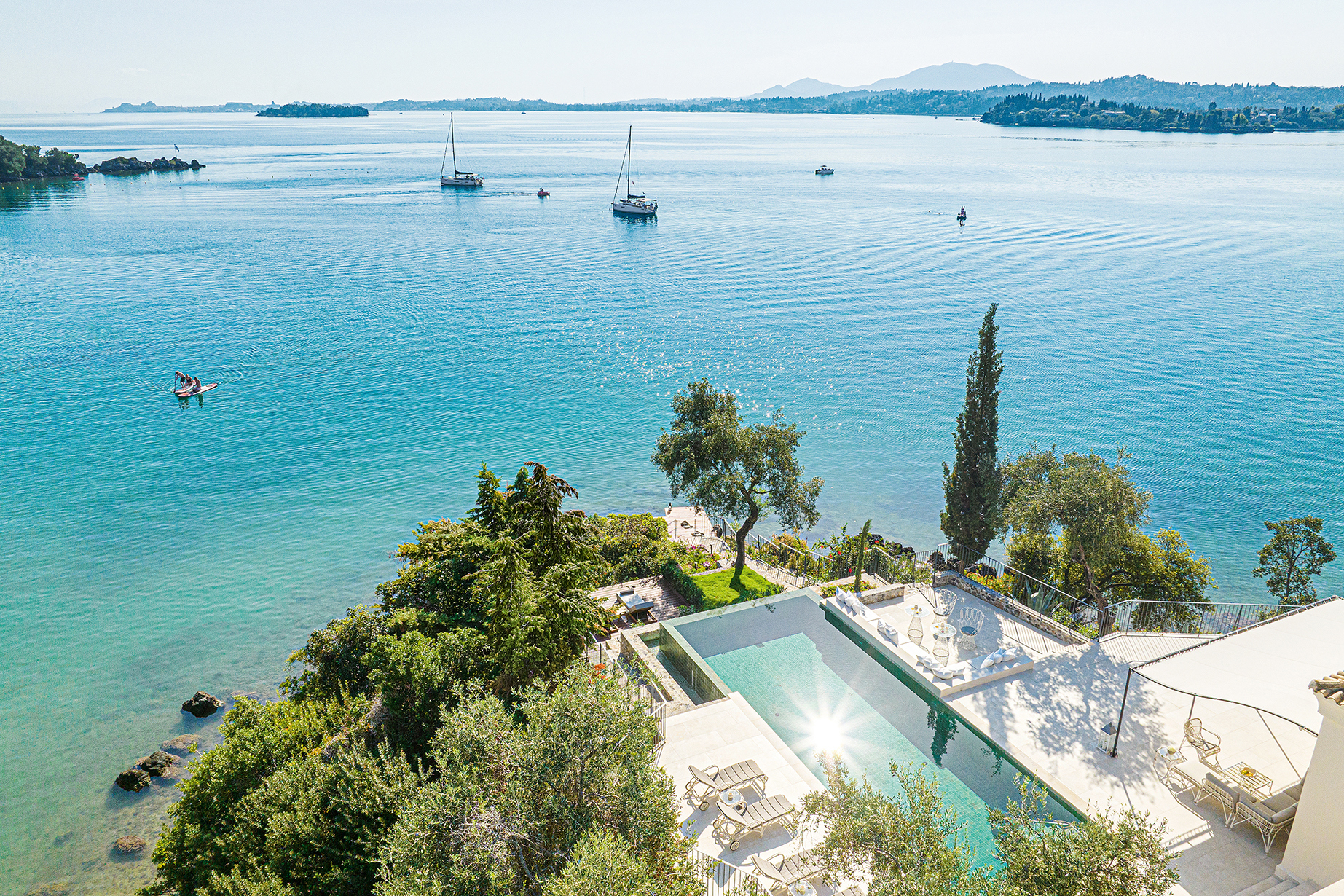 Aerial view of Grecotel Corfu Imperial’s Palazzo Imperiale pool overlooking the Ionian Sea with sailboats and lush coastal scenery.