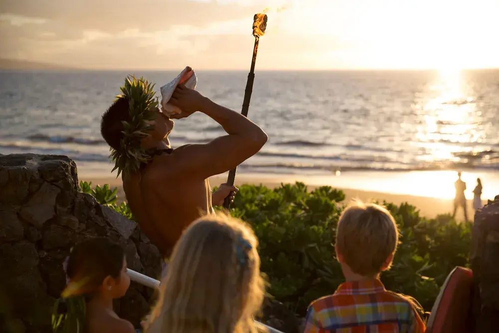 A person in traditional Hawaiian dress blowing into a conch shell, surrounded by young observers facing the ocean at sunset
