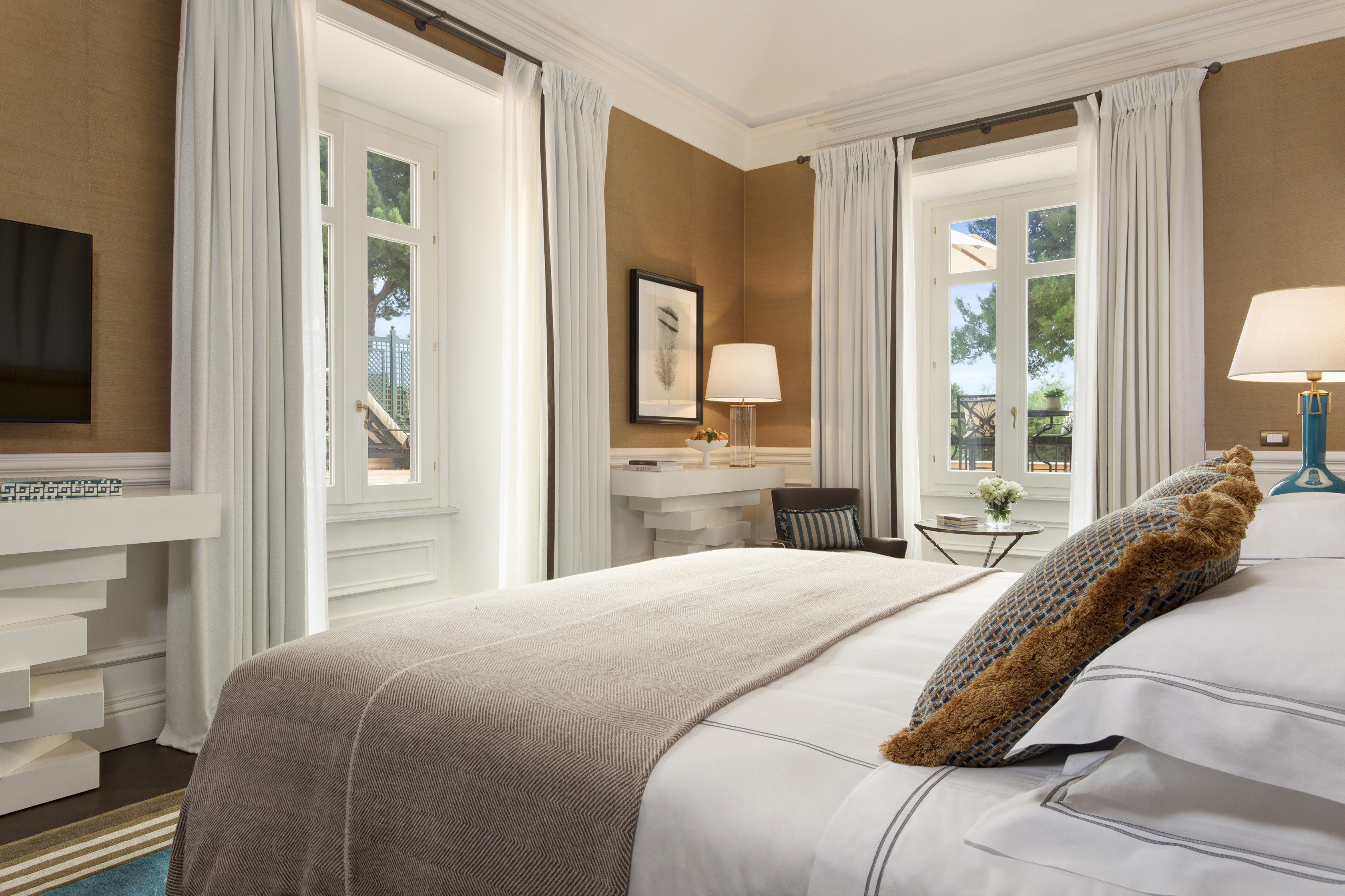 A well-lit, cozy bedroom featuring a large bed with beige and white bedding, a turquoise bedside lamp, white curtains, and a view of greenery outside the window.