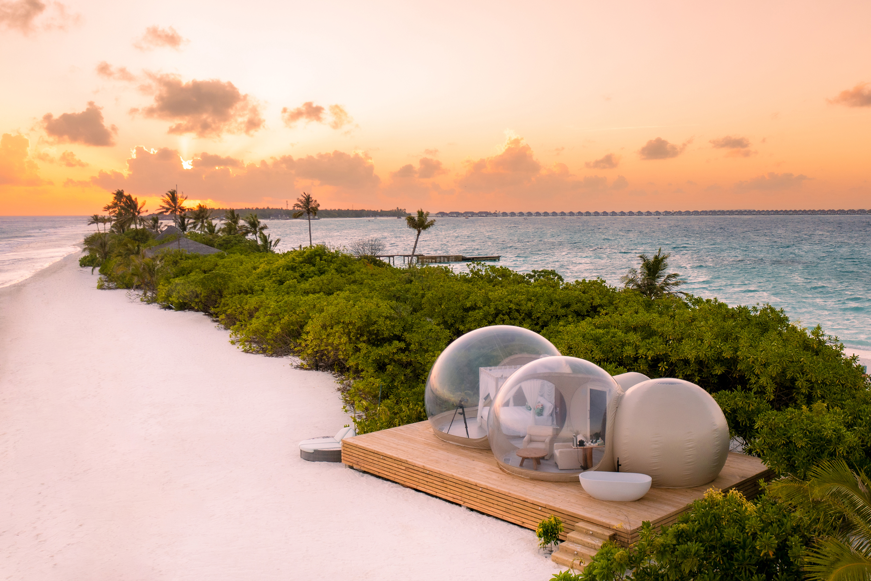 A private bubble suite on the sandbank of Finolhu with greenery stretching behind under a clear sky