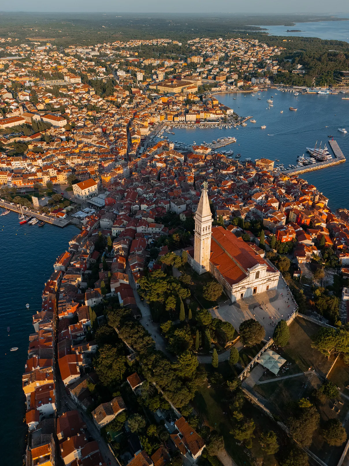 Aerial view of a historic coastal town with red-tiled rooftops, a prominent church and bell tower, and a harbor filled with boats along a blue waterfront.