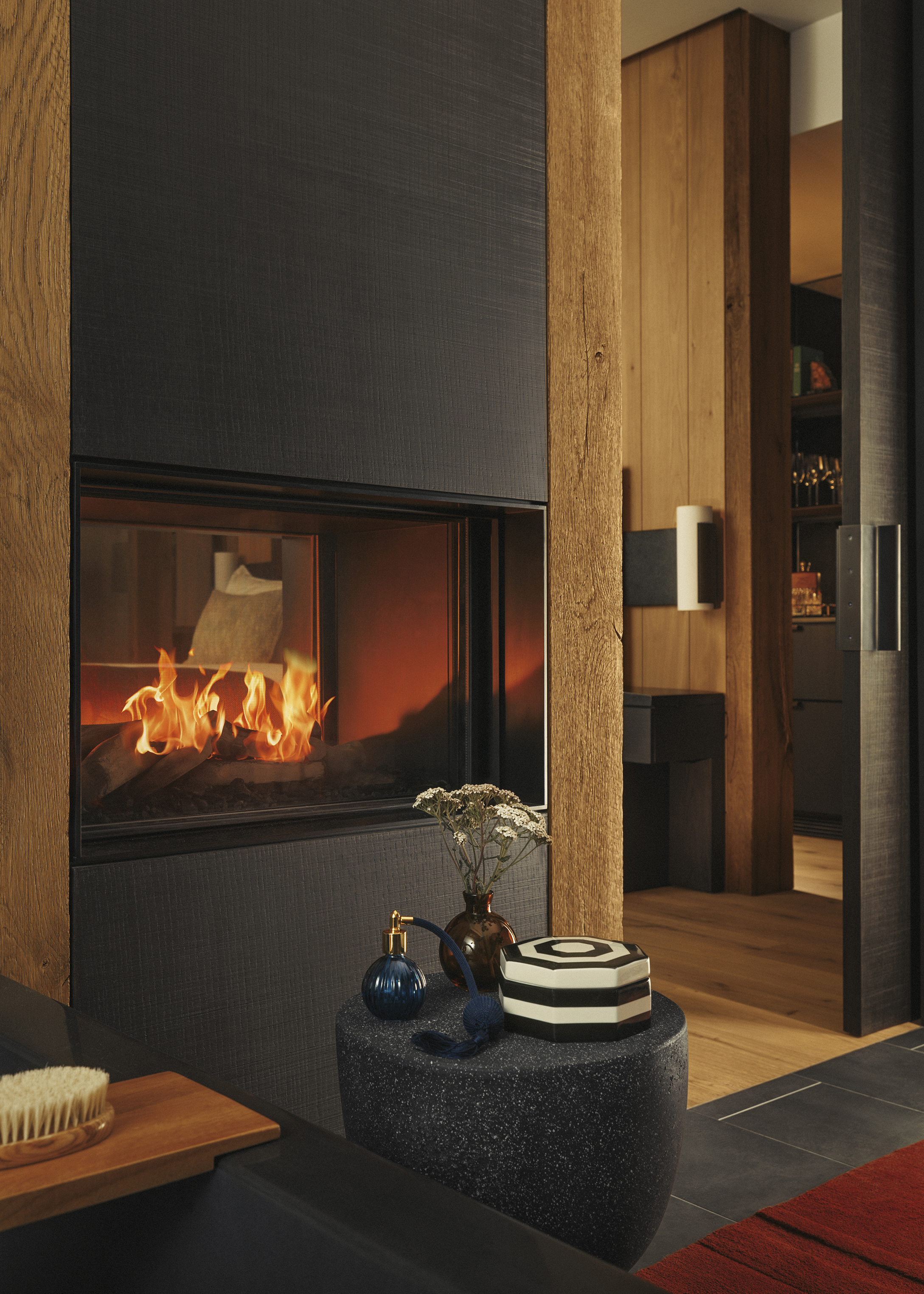 A sleek double sided fireplace with burning logs featuring a small table with a perfume bottle, ornamental box and vase on top