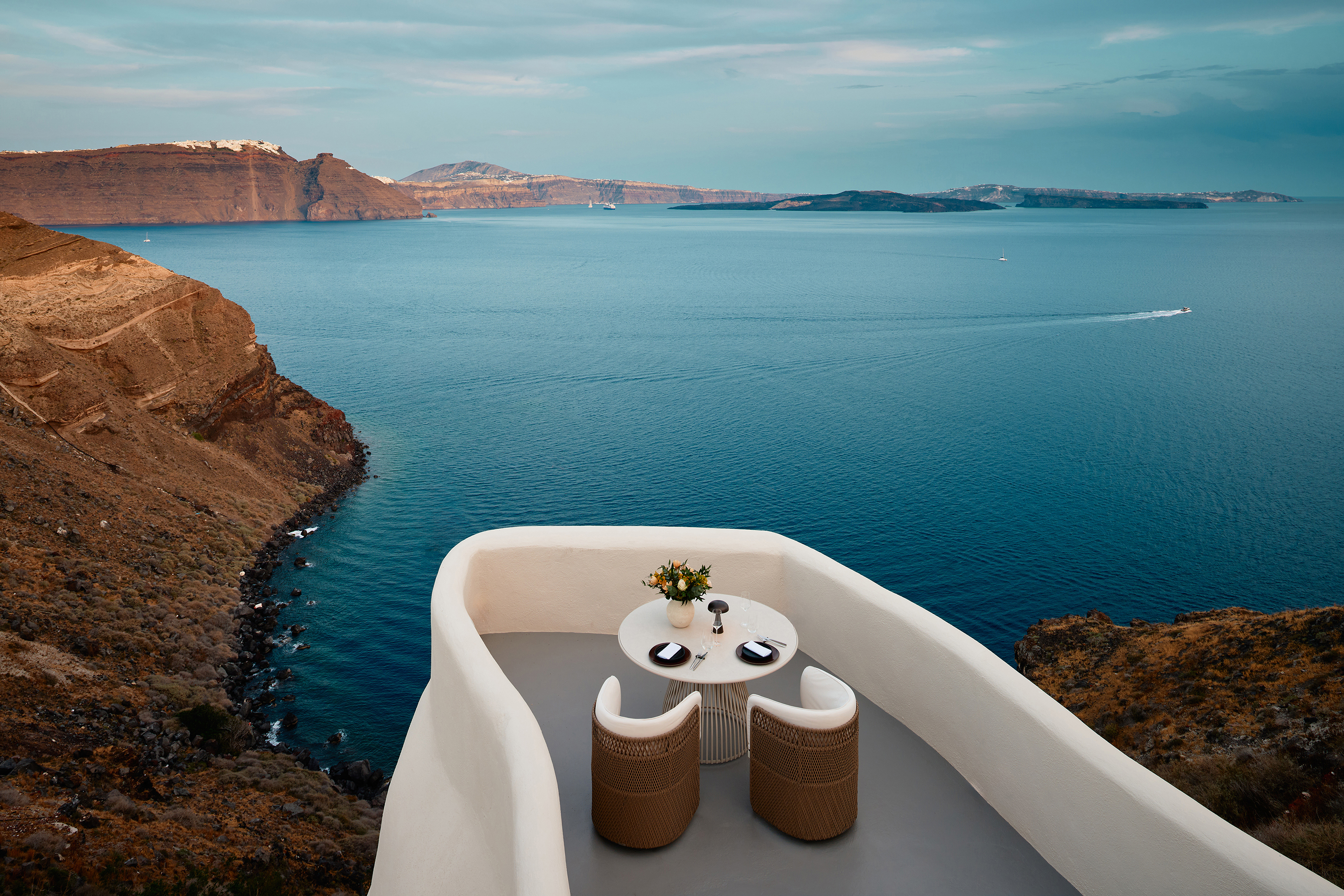 An intimate dining table set for a private meal at the end of a cliff front terrace overlooking a vast landscape with a boat on the sea