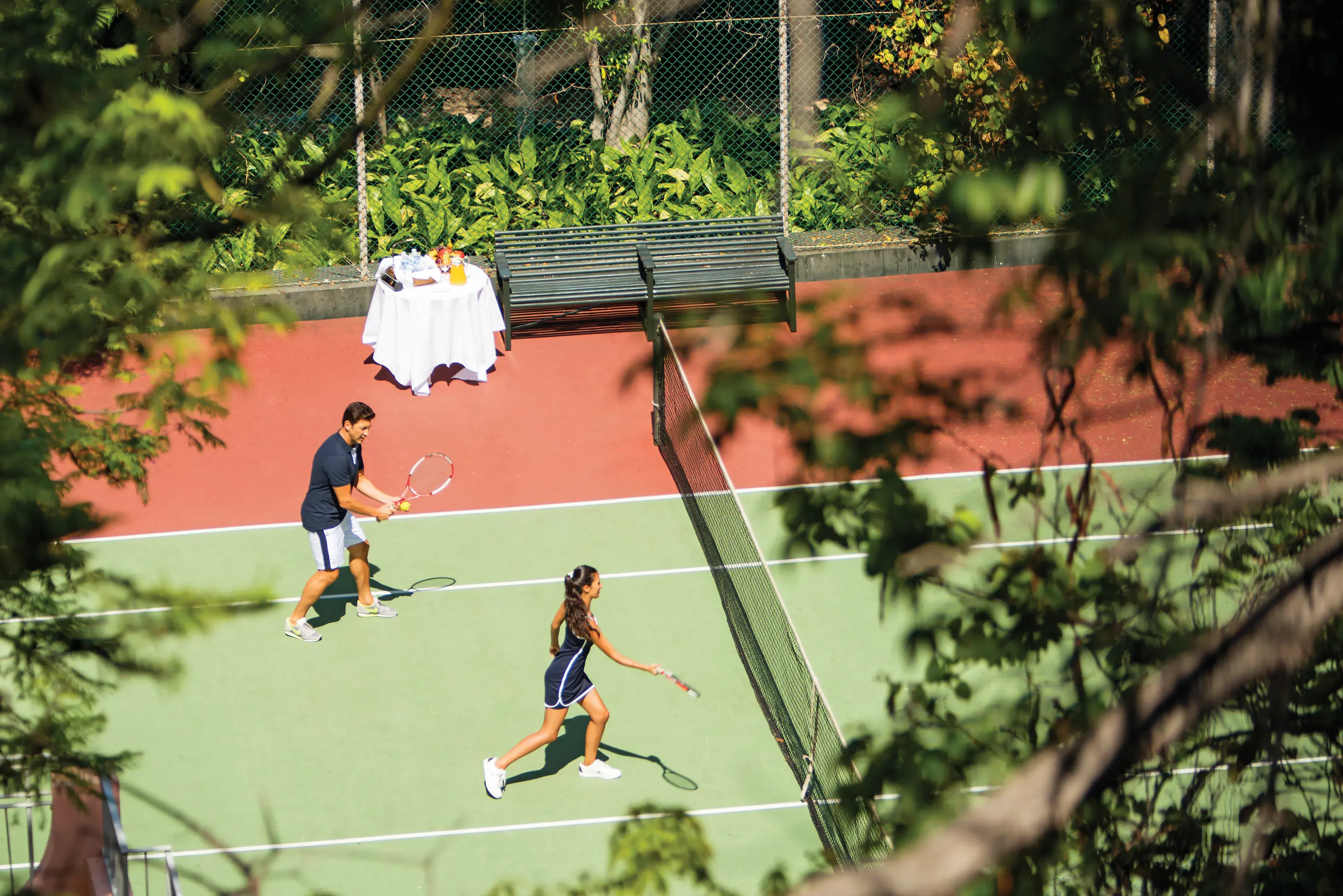 Two players rallying during a doubles tennis match on an outdoor hard court at Reid's Palace, A Belmond Hotel surrounded by lush greenery and trees.