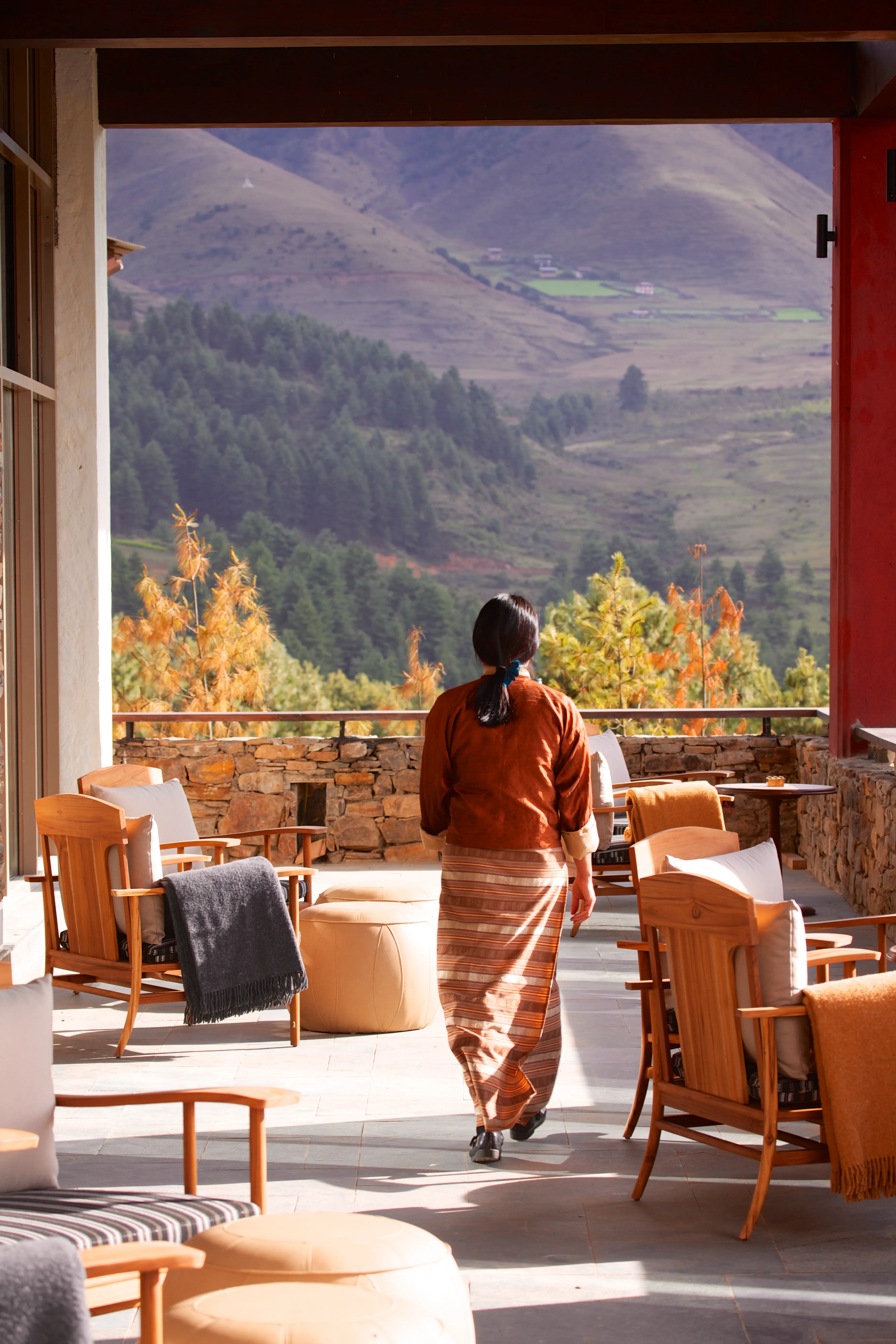 Staff member walking through a sunlit lodge terrace with wooden seating and panoramic mountain valley views.