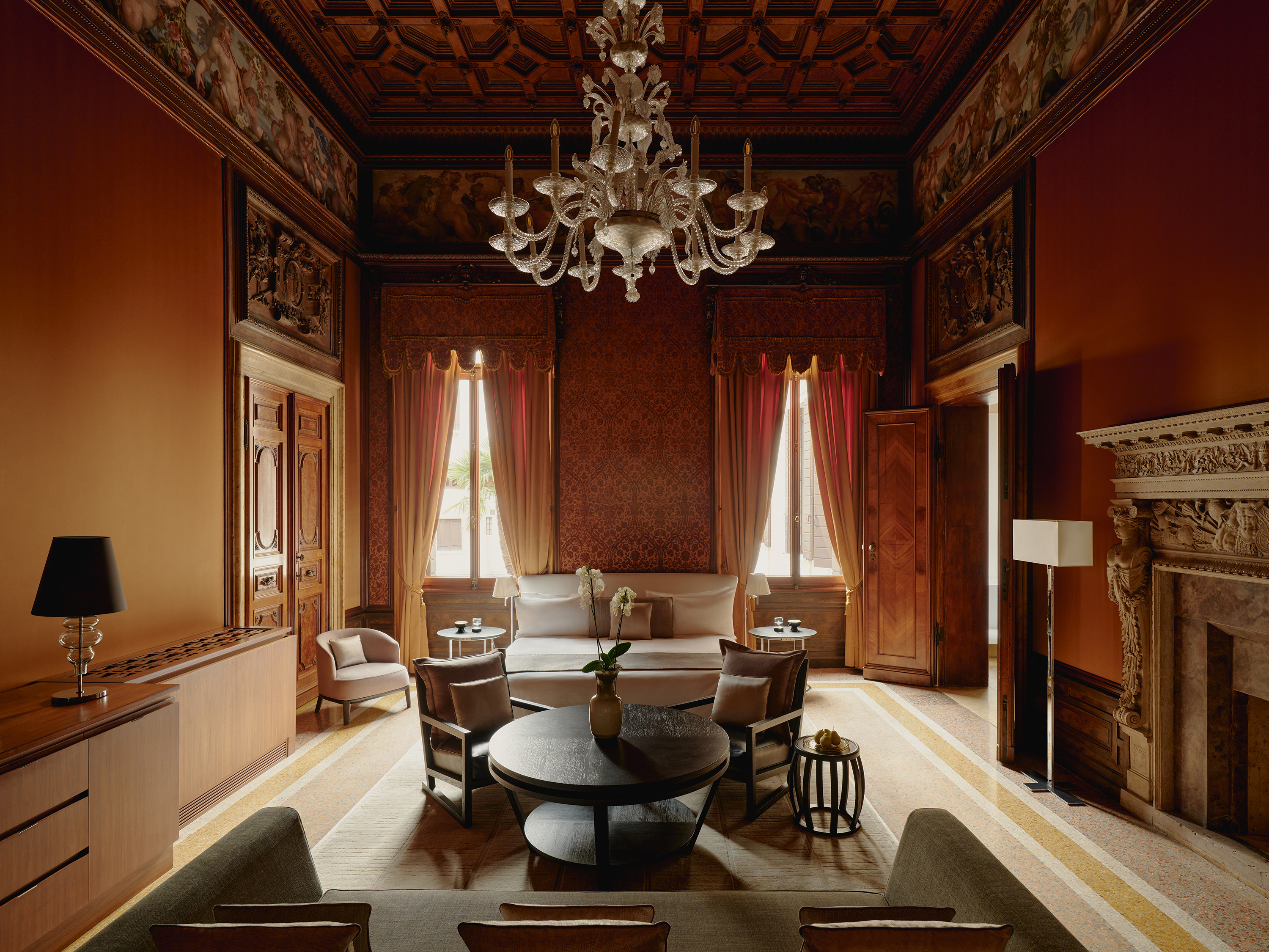 A lavish and moody bedroom with high ceilings decorated with patterns and paintings, featuring sleek contemporary furniture alongside an old fireplace and windows