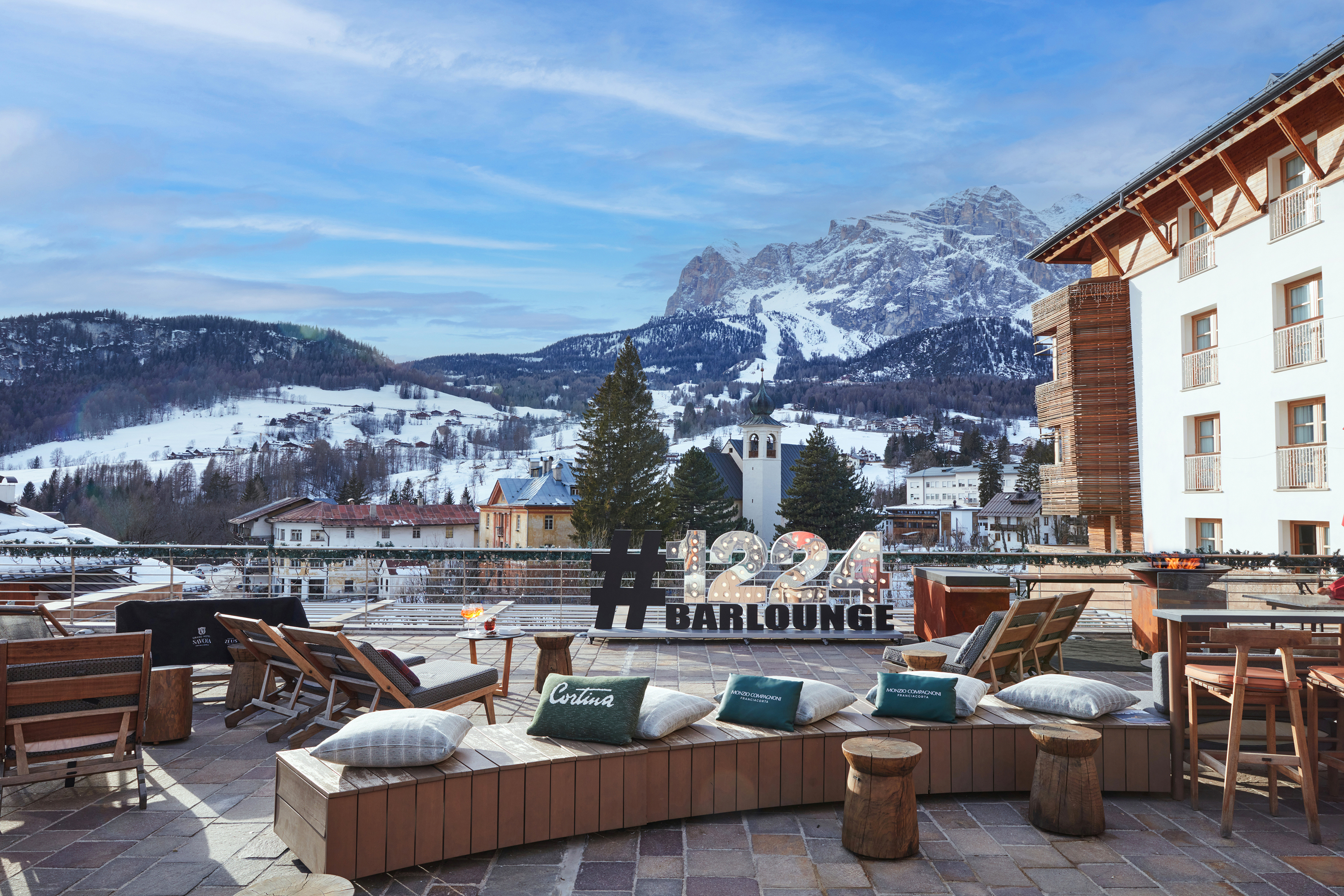 The open-air terrace of 1224 Bar at Grand Hotel Savoia with wooden outdoor furniture facing a sweeping view of the mountains beneath a clear sky