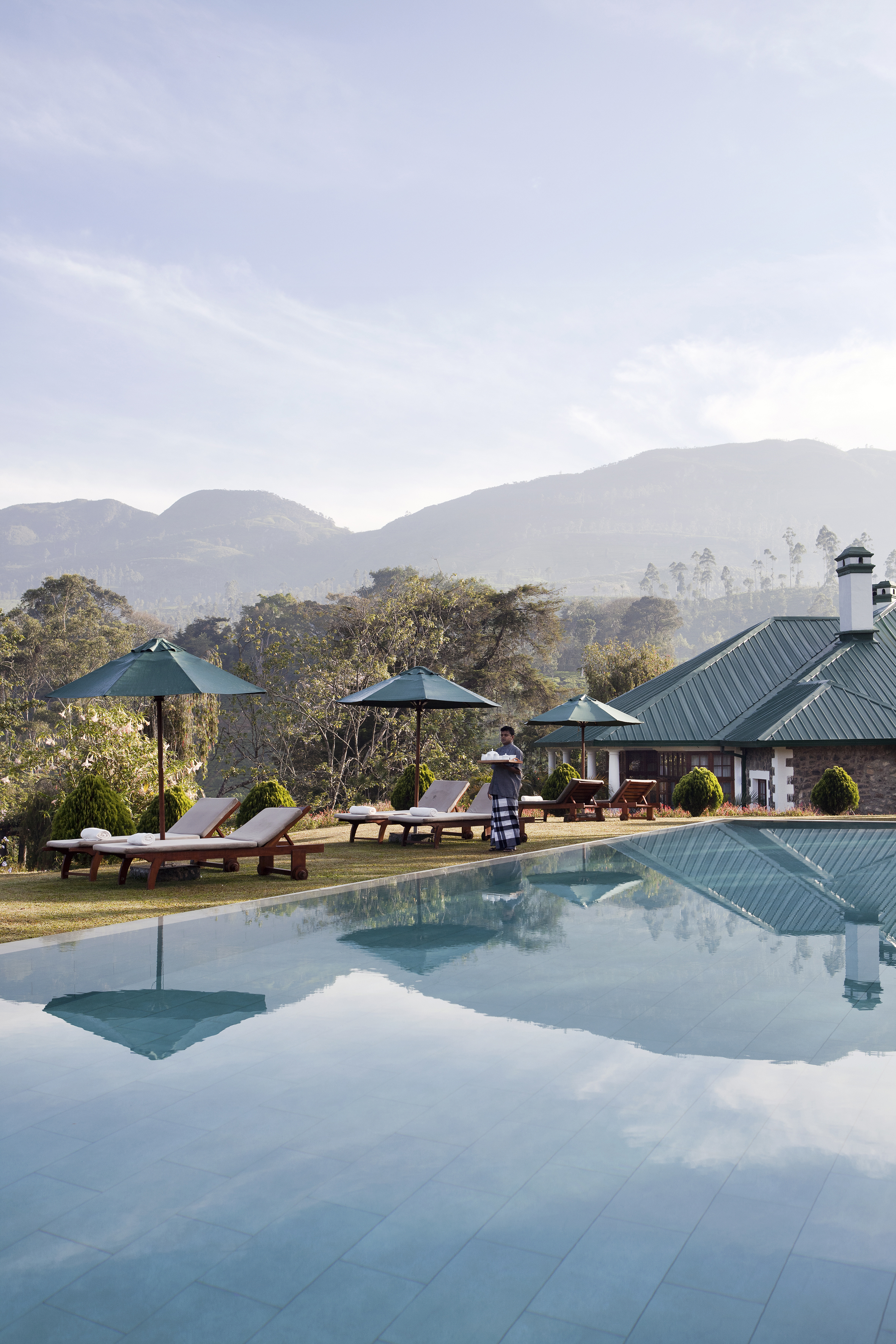 Man carrying a drinks tray walking beside a pool with sun loungers and hills in the background