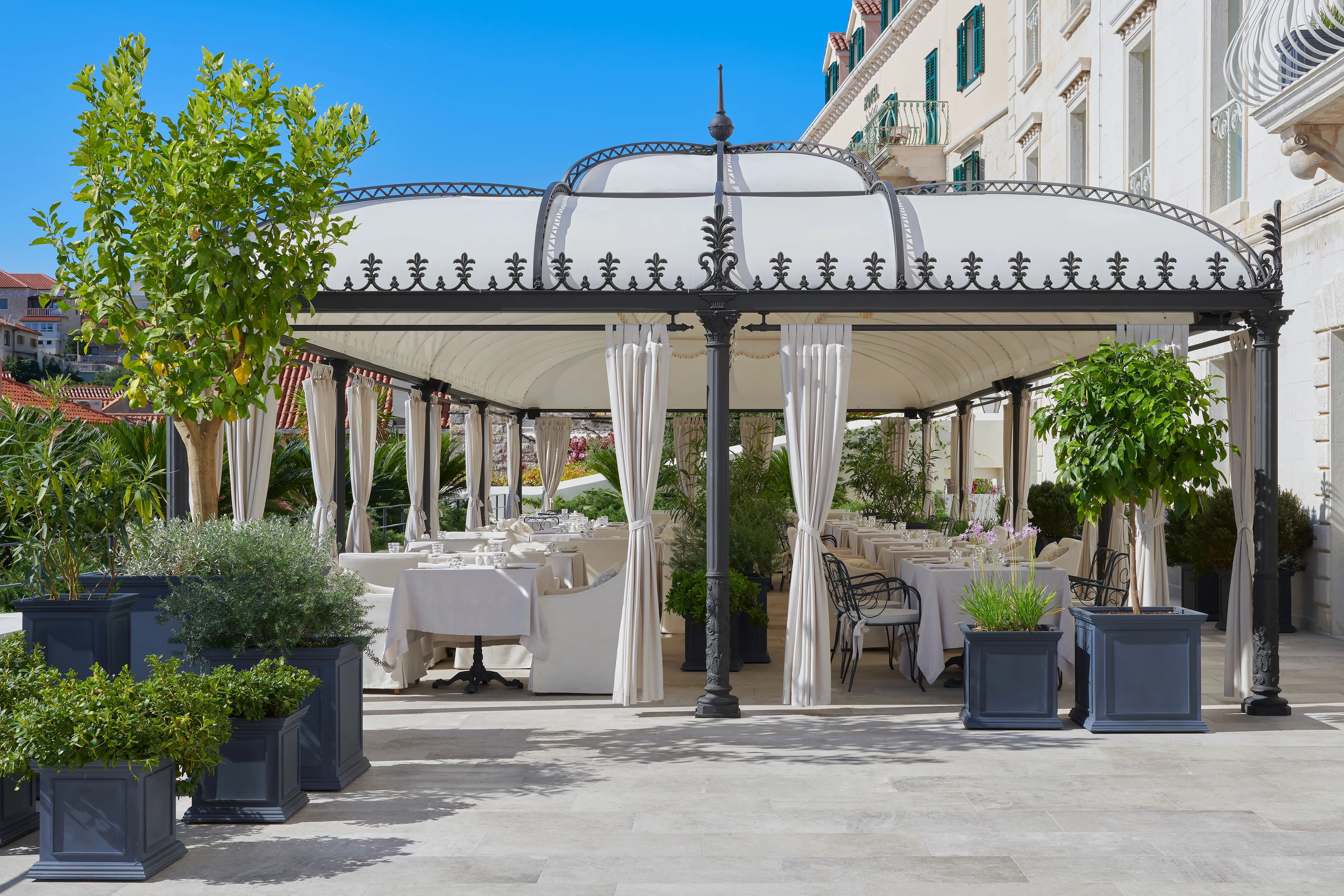 Terrace of San Marco Restaurant at Palace Elisabeth in Croatia featuring a Victorian-style cover with white dining tables beneath and potted plants