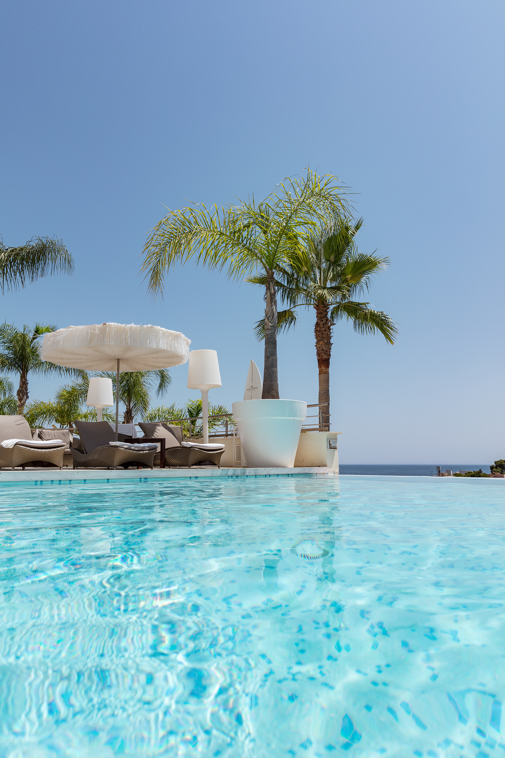Luxurious poolside at Portals Hills in Mallorca with clear blue water, palm trees, lounge chairs, and ocean views under a sunny sky.