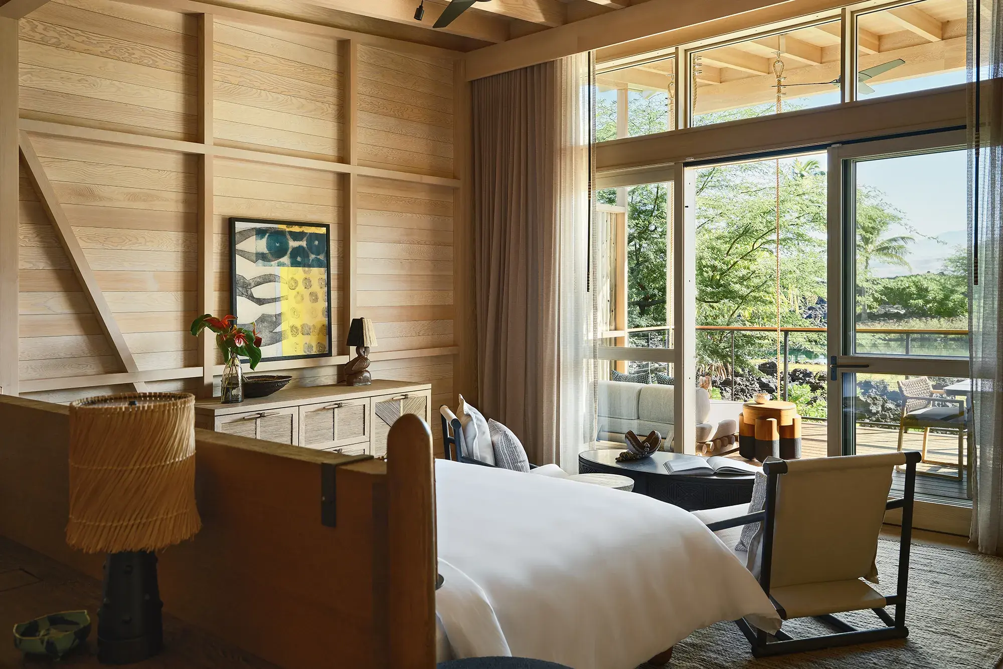 The bedroom of a Kauhale Mountain View featuring comfy wooden bed and furnishings with large glass doors and windows overlooking a terrace and lagoon