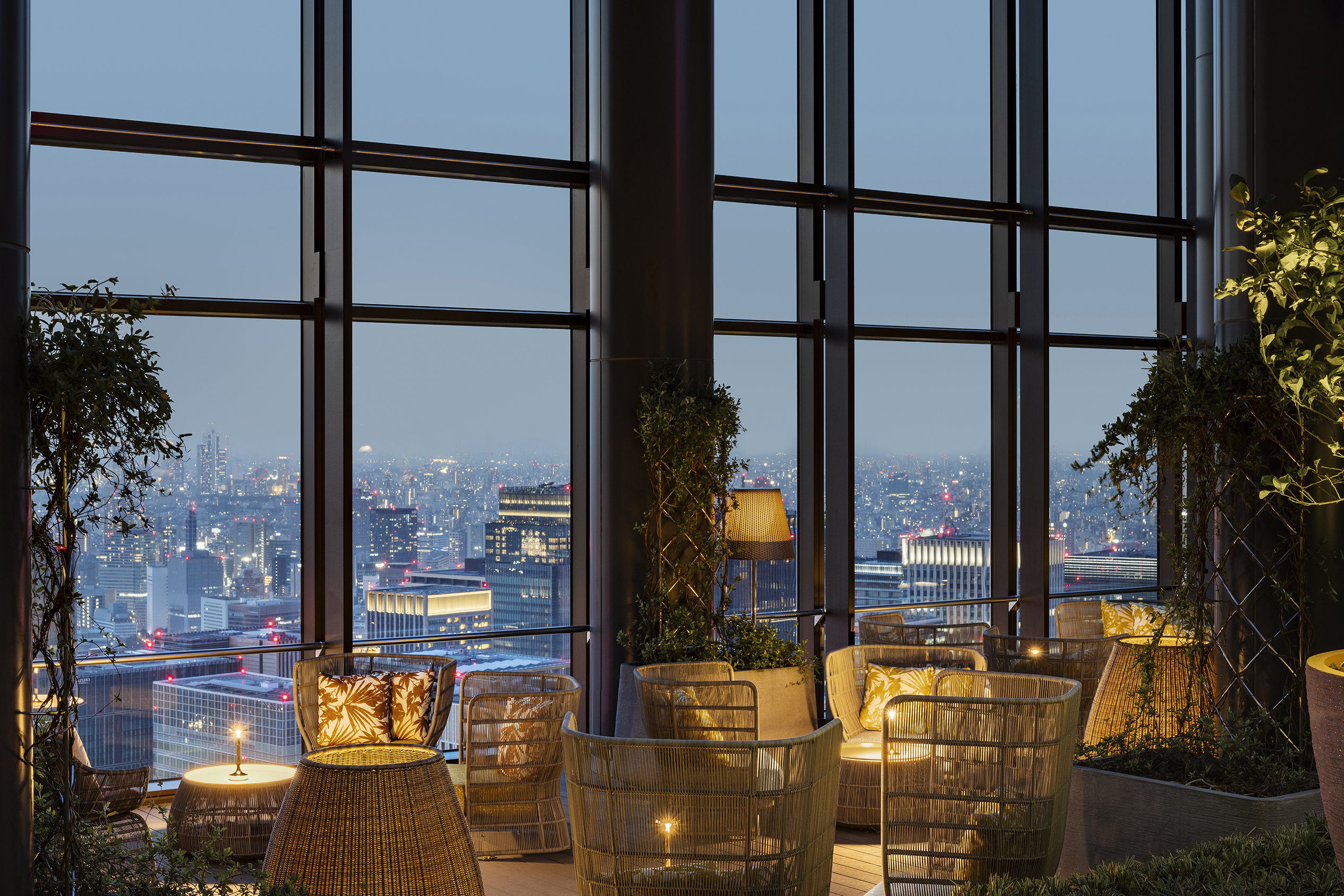 A lounge filled with rattan furniture and plants, with sweeping views of Tokyo lit up at dusk