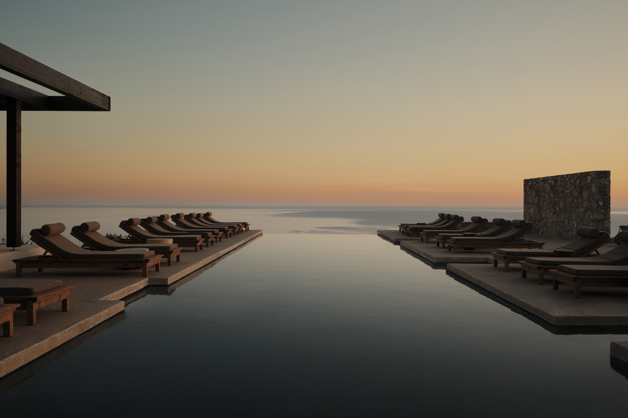 An infinity pool at sunset flanked by sun loungers reaching out towards the Agean sea