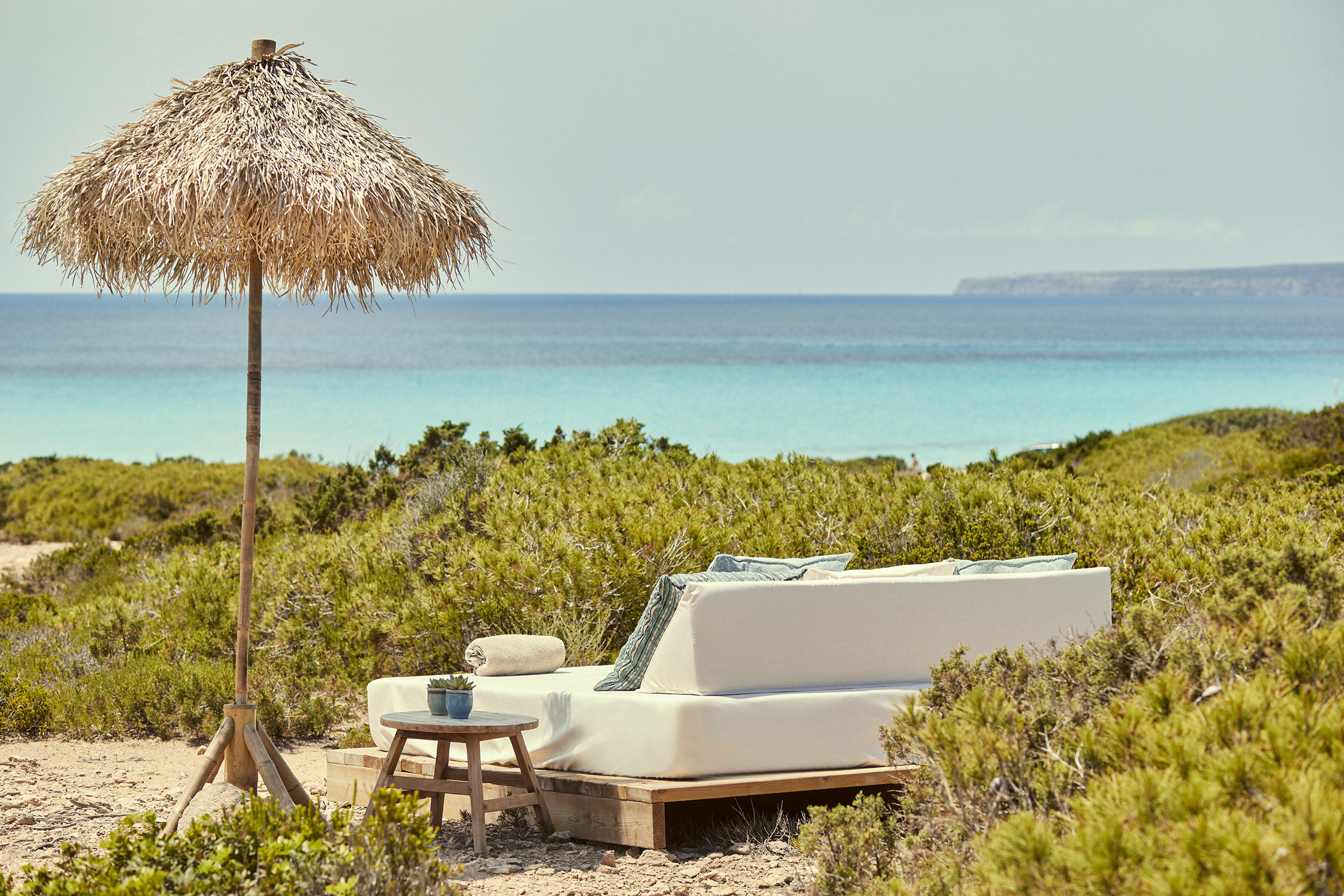 A comfy outdoor lounge area and straw umbrella at Teranka set beside coastal shrubbery with a view of the ocean under a clear sky