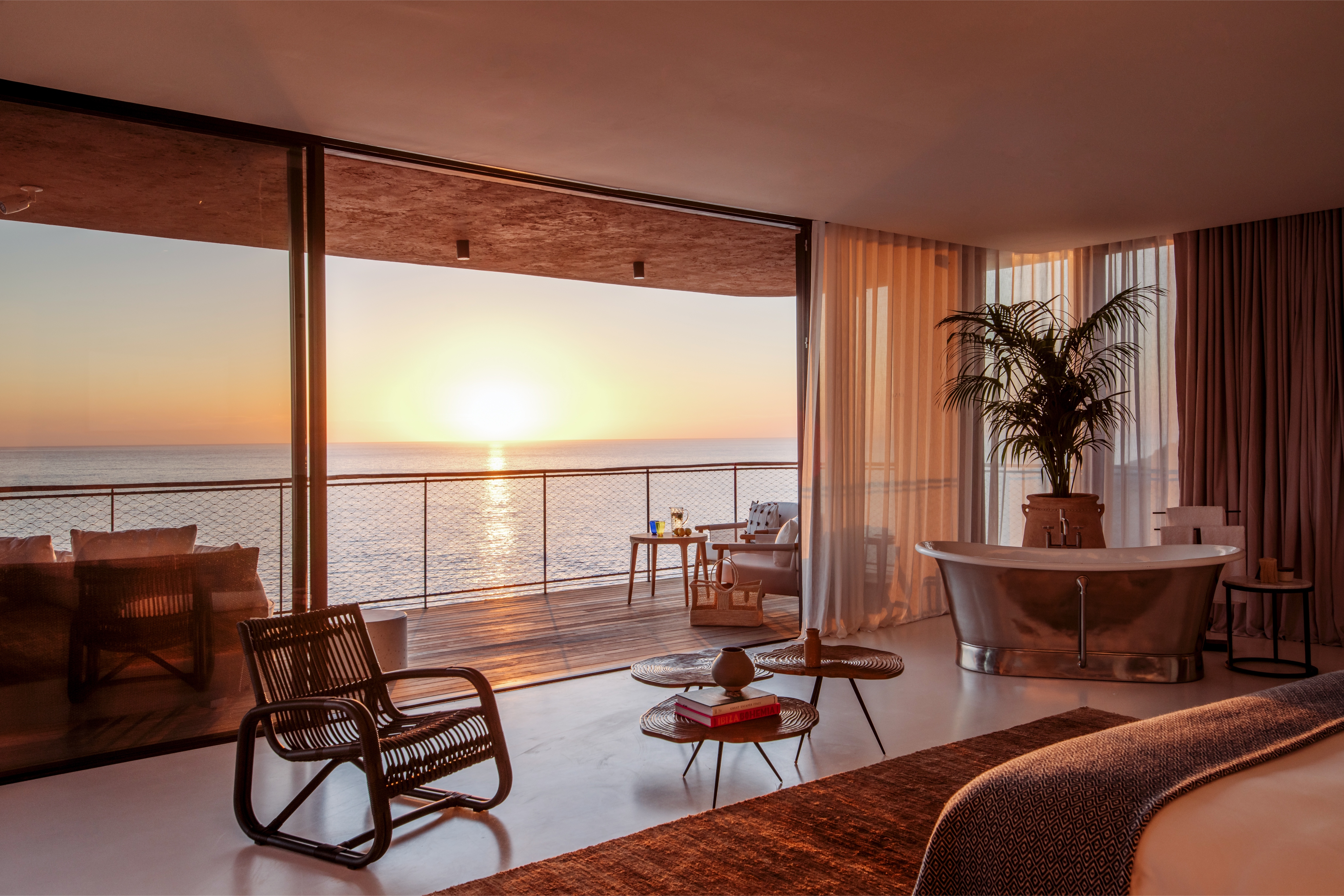 A luxurious bedroom with modern furniture and a large window overlooking an ocean sunset.