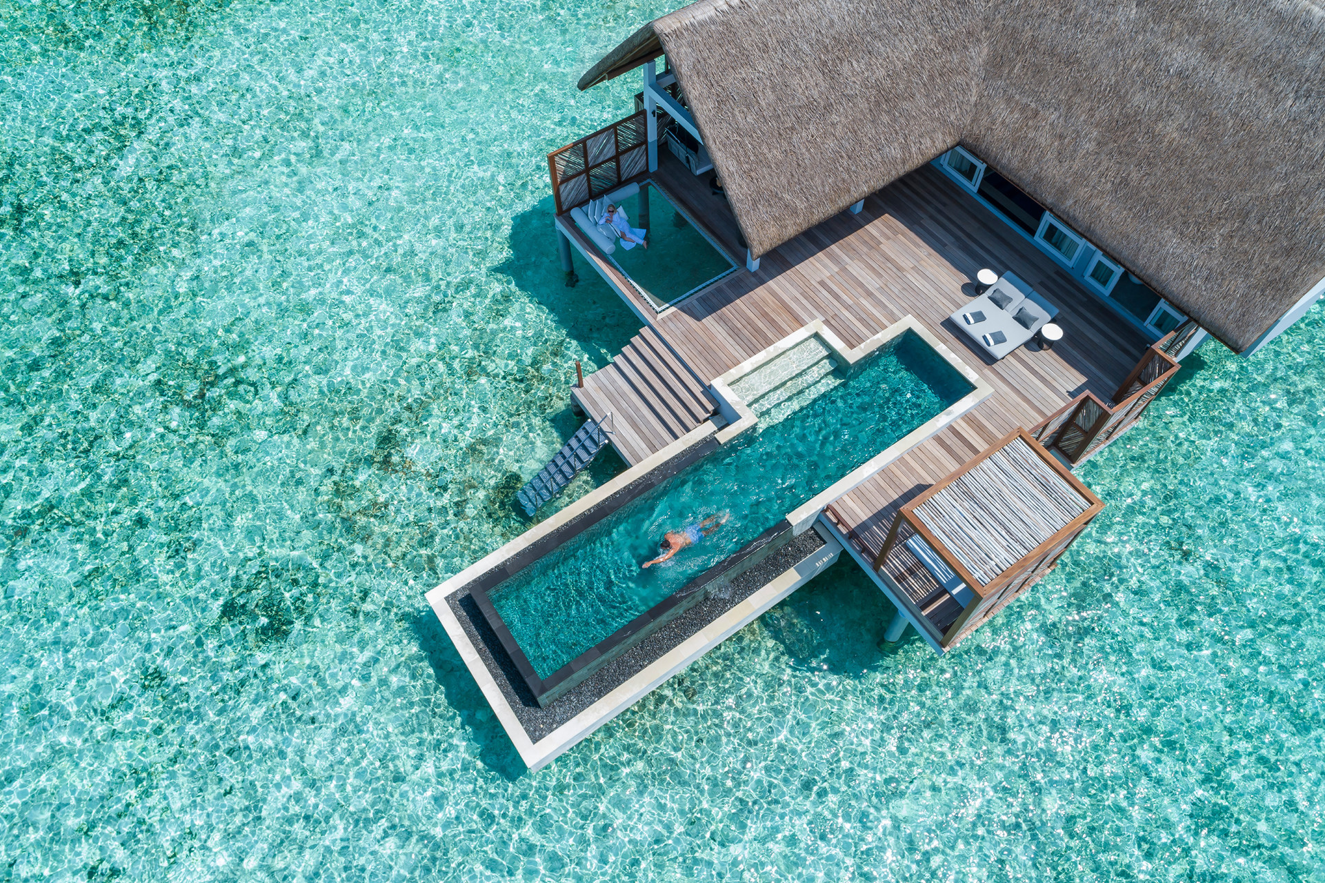 A birds eye view of an overwater villa with a person swimming in the private pool and person on a net lounge area