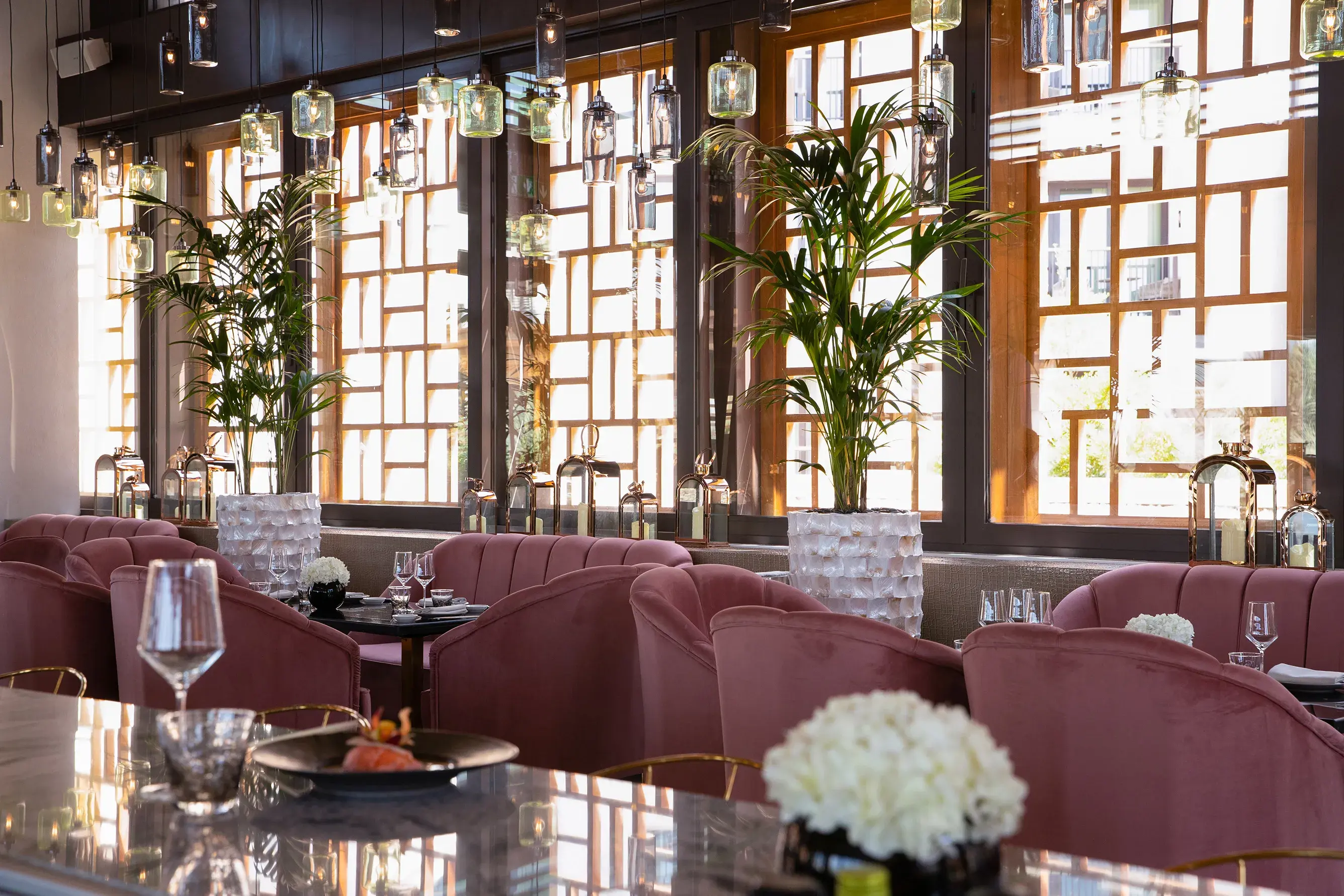 Elegant indoor dining space at Jumeirah Al Naseem Dubai featuring plush pink seating, marble tables, hanging glass lights and tall potted palms.